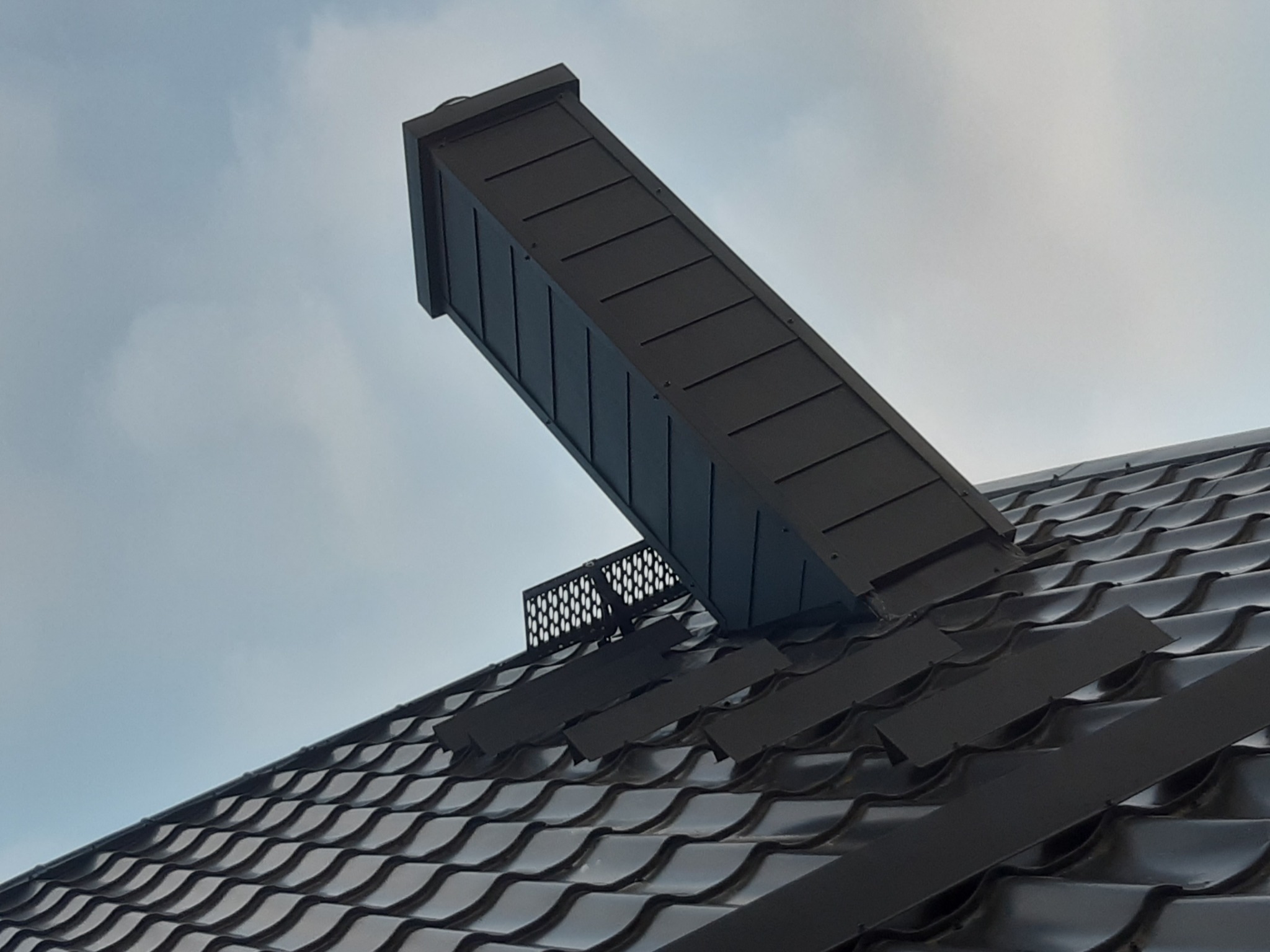 Nisko kątowe ujęcie ciemnej, błyszczącej dachówki ceramicznej z zamontowanym, prostokątnym kominkiem wentylacyjnym z metalową kratką wentylacyjną, na tle pochmurnego nieba.