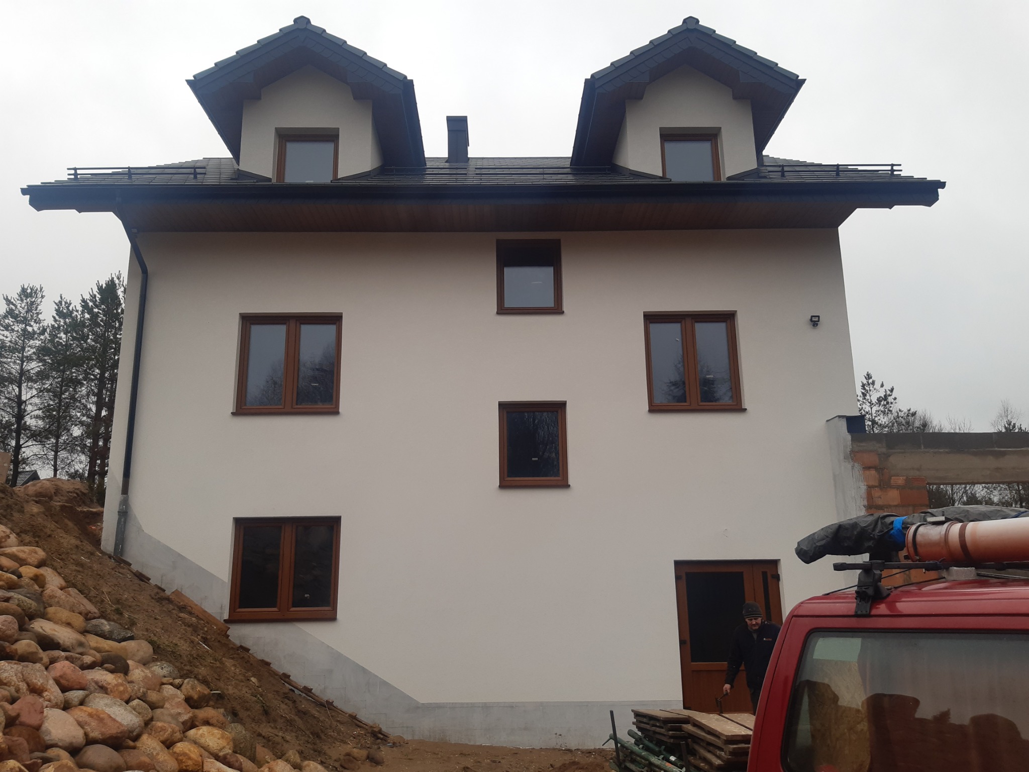 Dwukondygnacyjny dom z jasną elewacją i ciemnym dachem, widoczny z zewnątrz, z elementami rusztowania i samochodem dostawczym w kadrze, sugerujący trwające prace wykończeniowe.