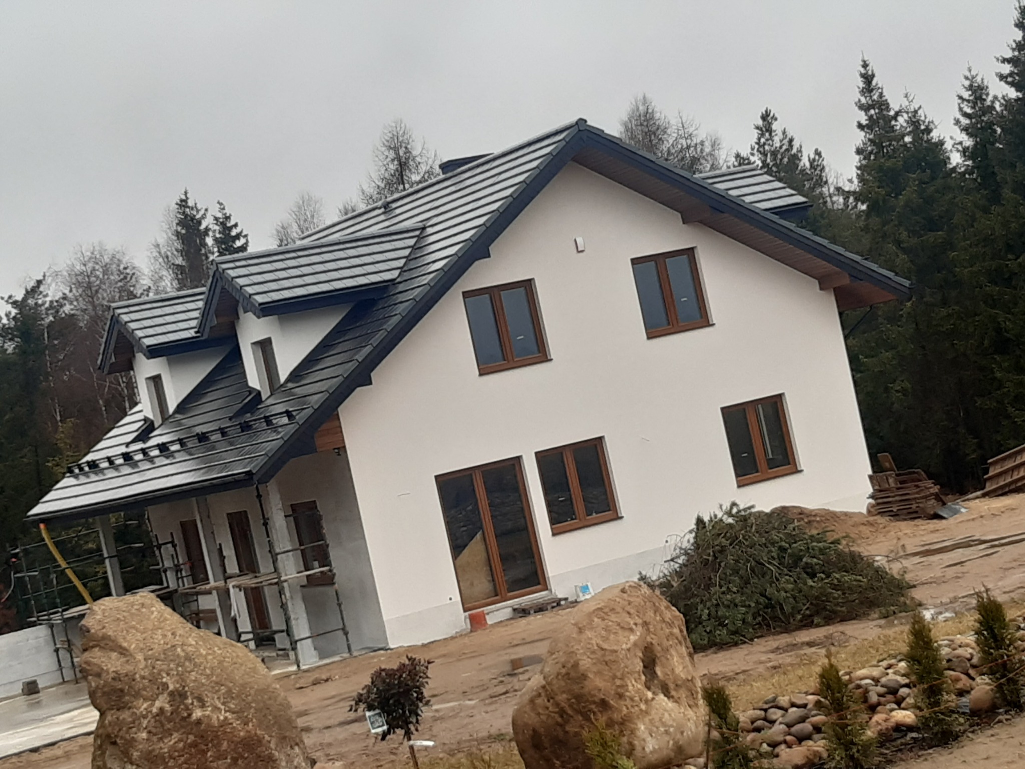 Nowoczesny dom jednorodzinny z ciemną dachówką i białymi ścianami, widoczne rusztowania i elementy wykończeniowe elewacji, teren w trakcie zagospodarowania, duże kamienie ozdobne na pierwszym planie.