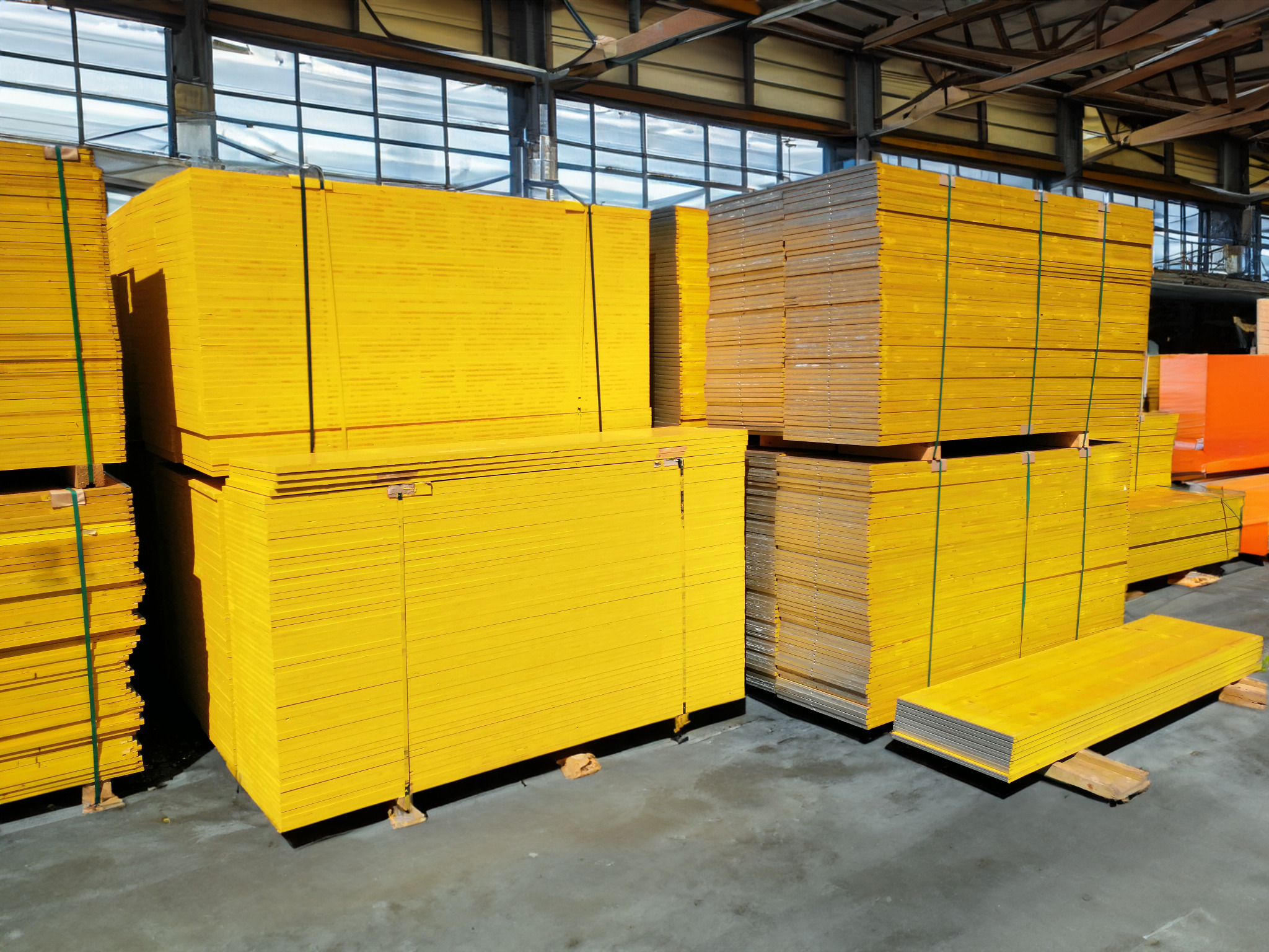 Magazynowane żółte płyty szalunkowe ułożone w równe stosy, zabezpieczone zieloną taśmą, widoczne w hali z oknami dachowymi.
