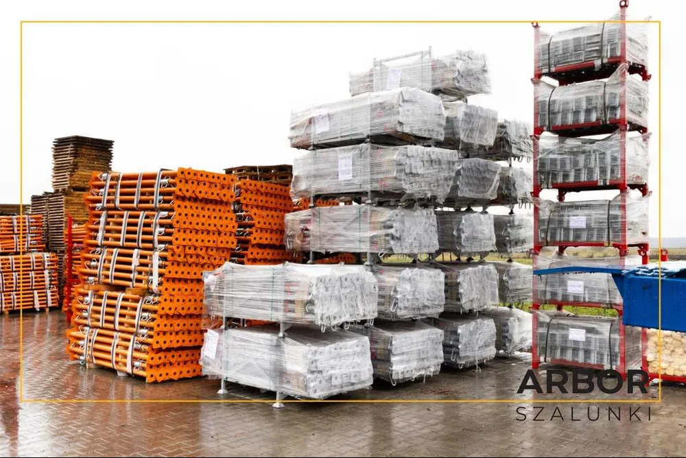 Sztaple nowych, zapakowanych w folię szalunków stalowych i pomarańczowych podpór budowlanych, gotowych do transportu, na mokrym placu.