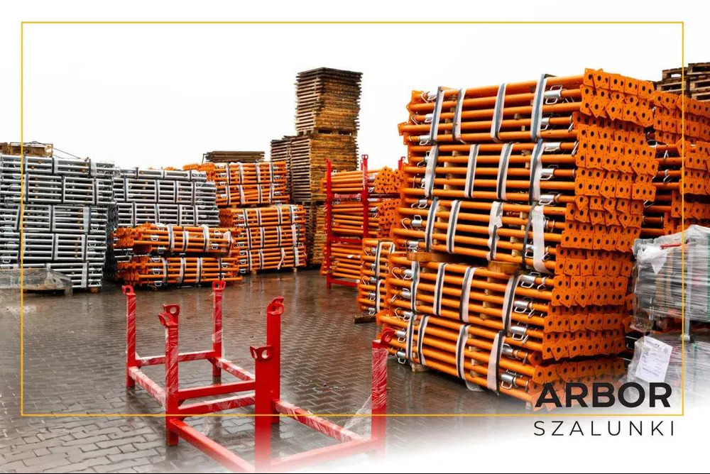 Sztaple pomarańczowych i szarych szalunków budowlanych na mokrym placu składowym, z czerwonymi ramami transportowymi na pierwszym planie.