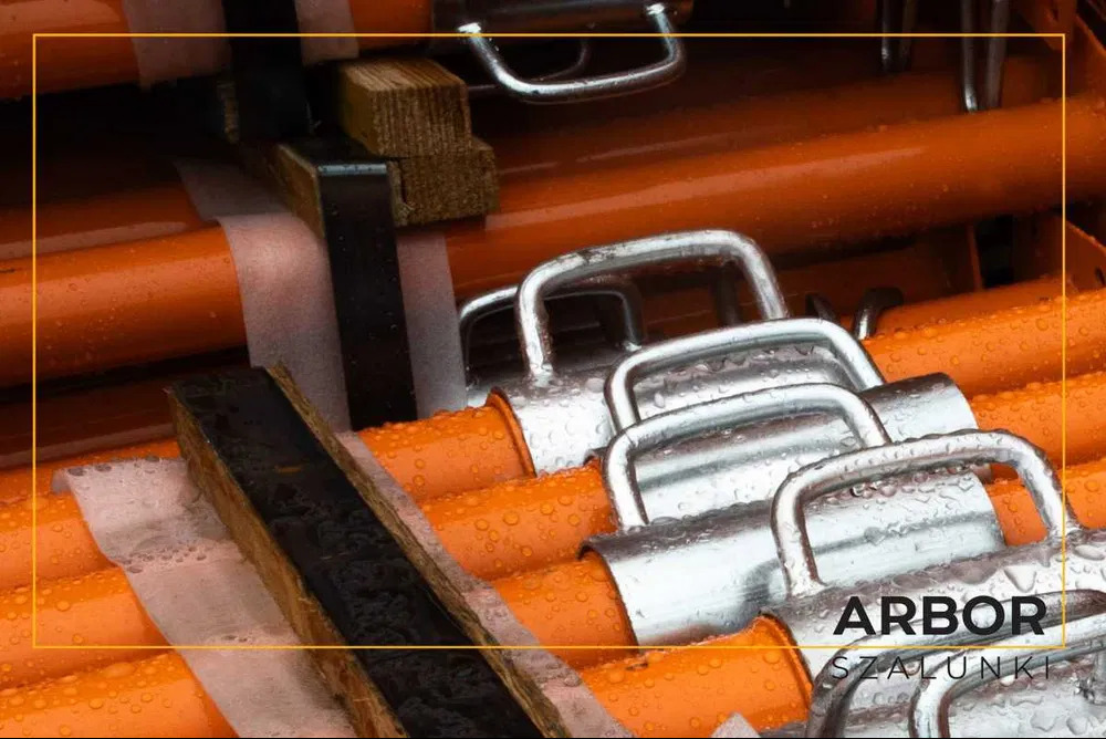 Pomarańczowe podpory szalunkowe z metalowymi uchwytami, zabezpieczone folią, ułożone warstwami, widoczne krople wody.