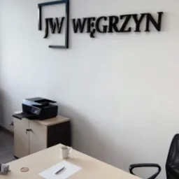 Biuro doradcy z logo firmy 'JW Węgrzyn' na ścianie, biurko z laptopem, kartką i długopisem, filiżanka kawy oraz drukarka na szafce.