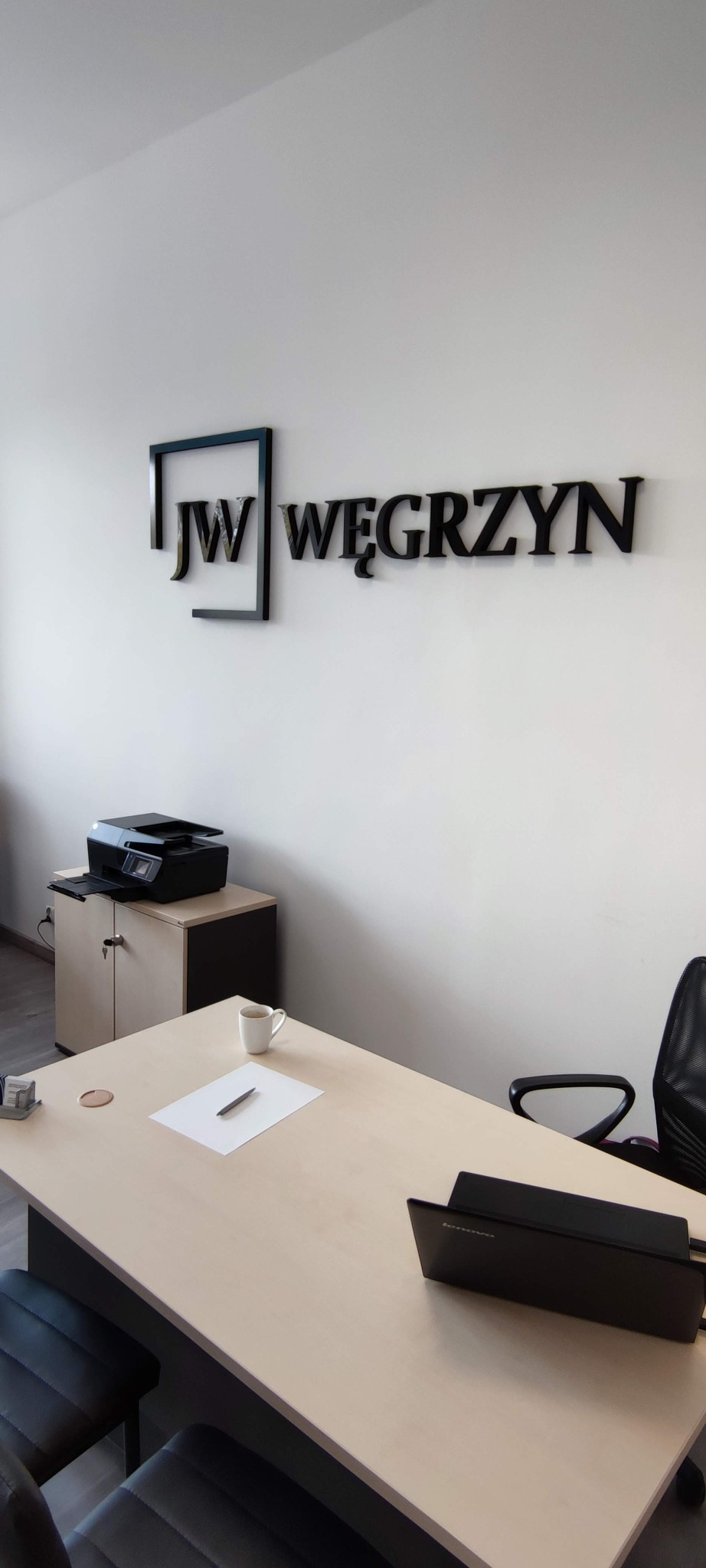 Biuro doradcy z logo firmy 'JW Węgrzyn' na ścianie, biurko z laptopem, kartką i długopisem, filiżanka kawy oraz drukarka na szafce.