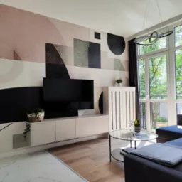 Jasne wnętrze salonu z geometryczną tapetą w odcieniach różu, szarości i czerni, minimalistyczną szafką RTV, okrągłym szklanym stolikiem kawowym z butelką wina i kieliszkami, oraz szarą sofą narożną.