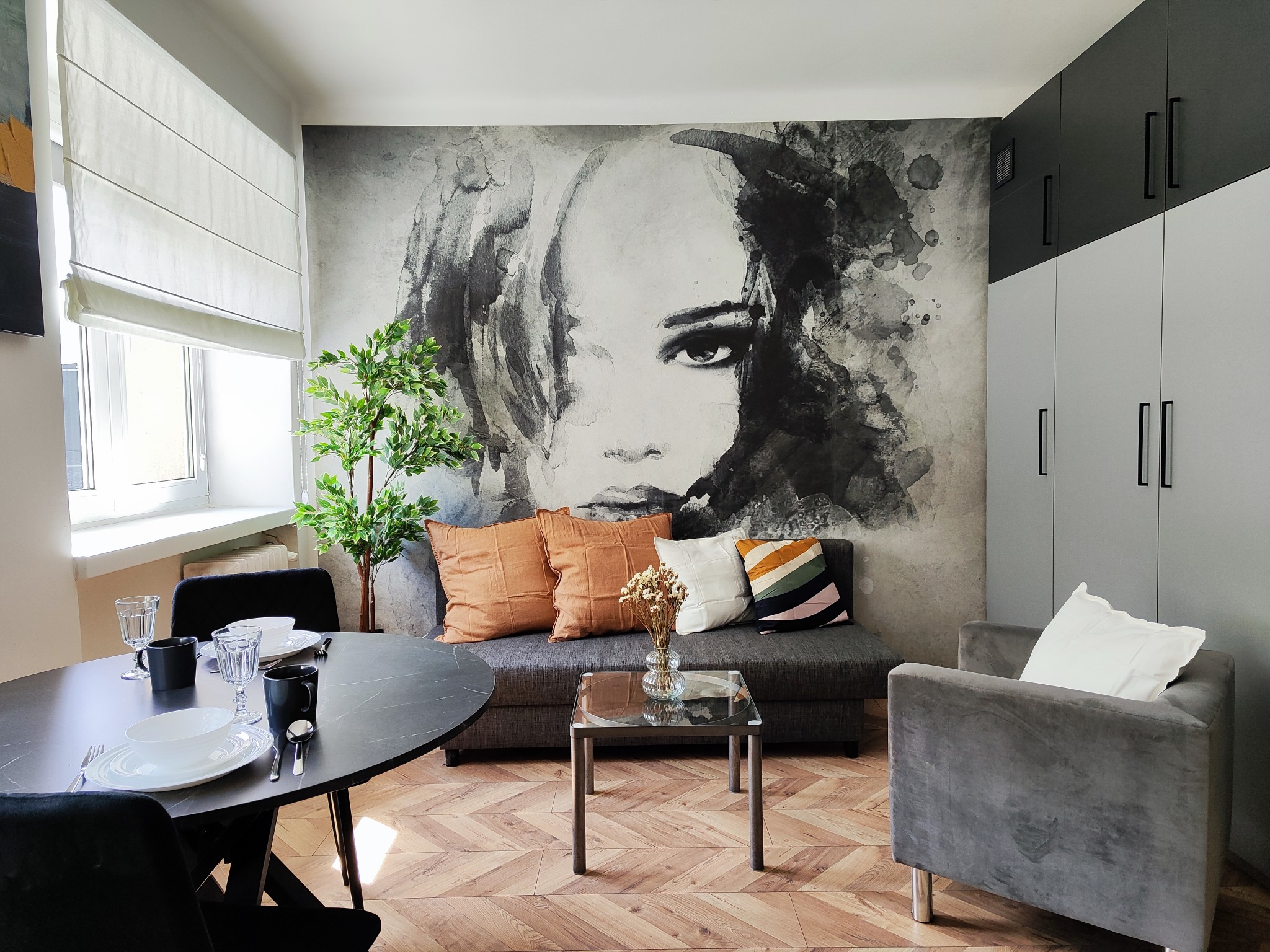 Jasne wnętrze salonu z ciemnym okrągłym stołem, szarą sofą z dekoracyjnymi poduszkami, szarym fotelem i tapetą z motywem portretu kobiety.