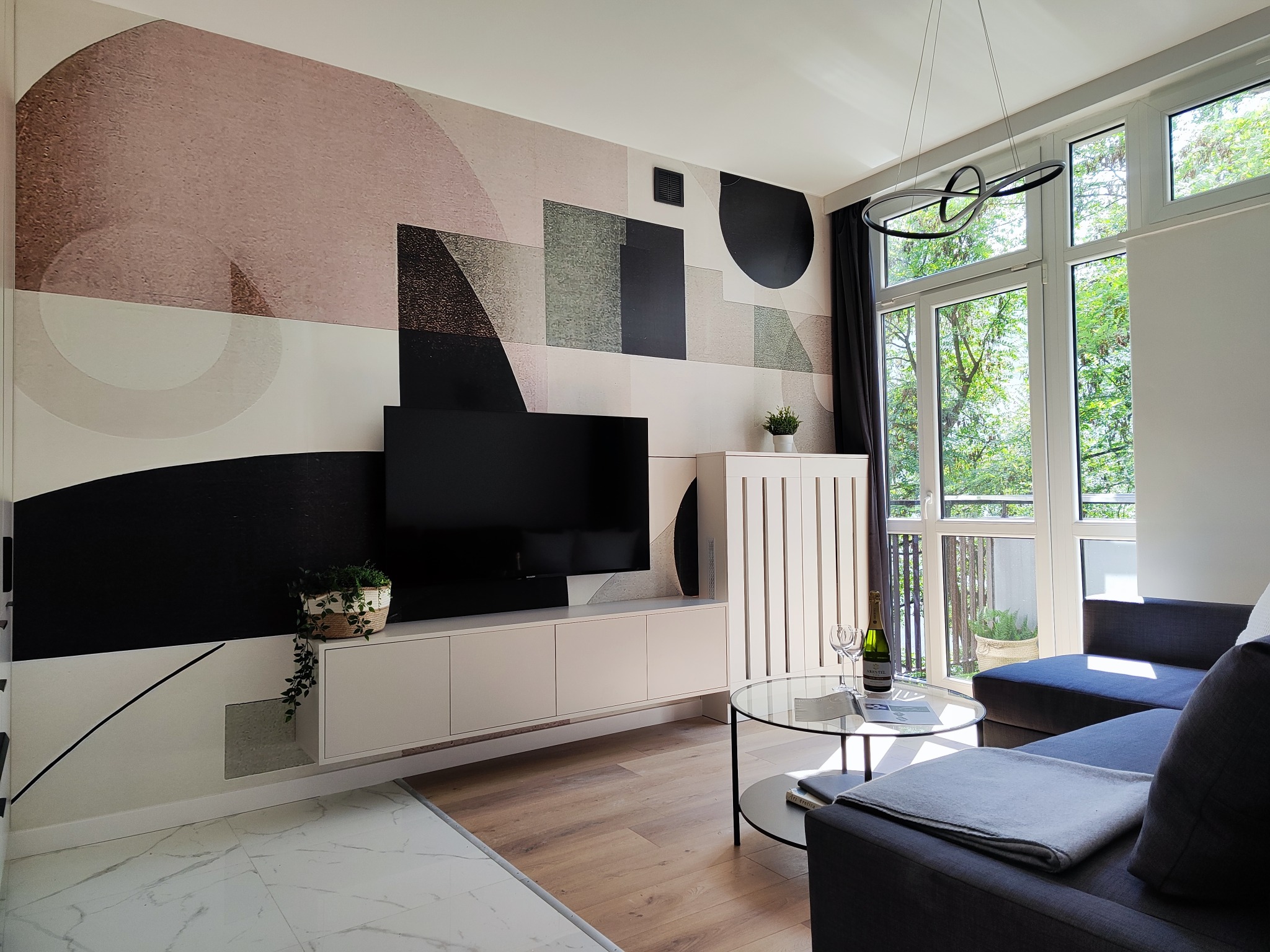 Jasne wnętrze salonu z geometryczną tapetą w odcieniach różu, szarości i czerni, minimalistyczną szafką RTV, okrągłym szklanym stolikiem kawowym z butelką wina i kieliszkami, oraz szarą sofą narożną.