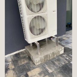 WK INSTALACJE SPÓŁKA Z OGRANICZONĄ ODPOWIEDZIALNOŚCIĄ - Zewnętrzna jednostka klimatyzacji dwuwentylatorowa na betonowym cokole z widocznymi śladami wilgoci, zamontowana przy ciemnoszarym budynku, z butelką pod jednostką.