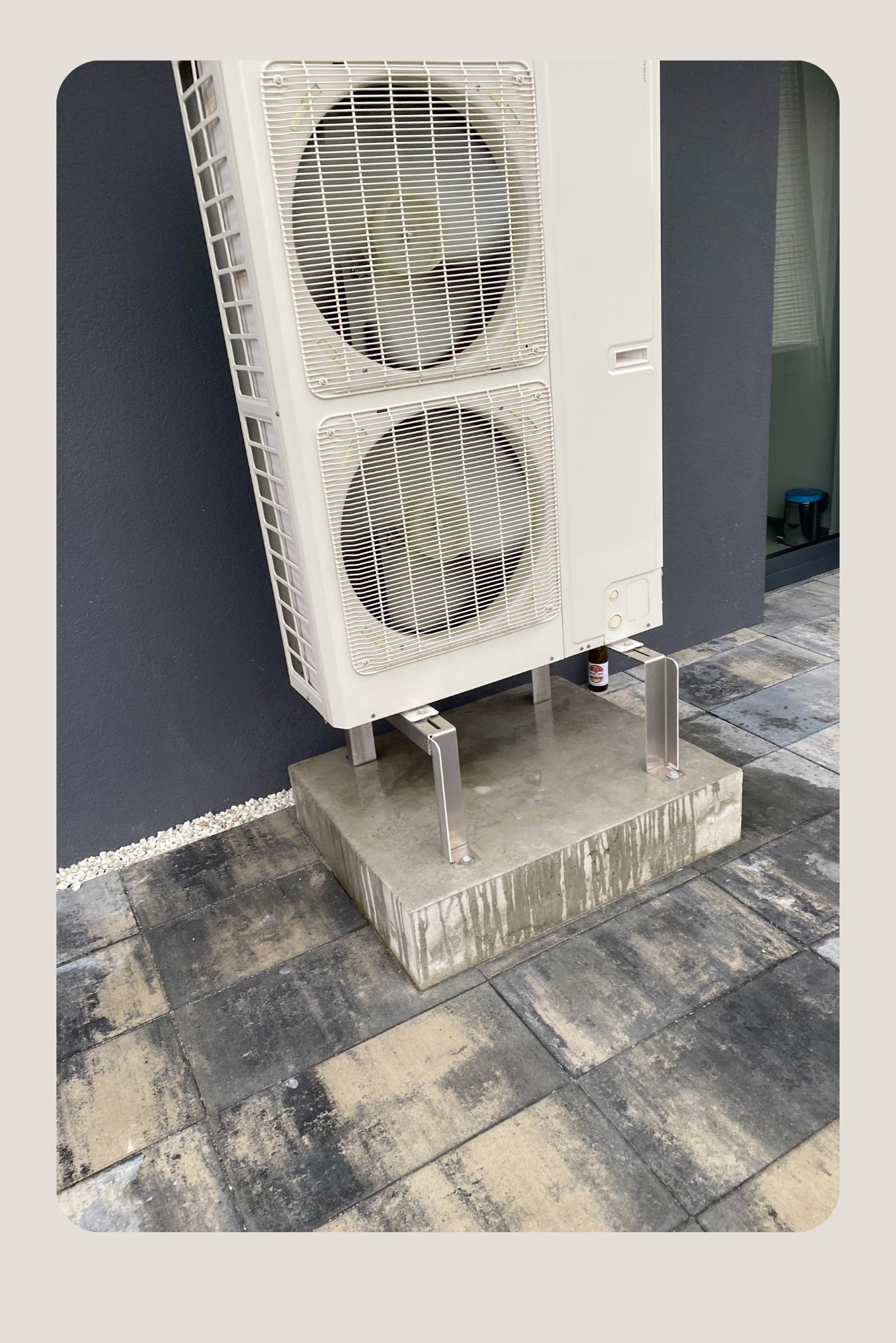 Zewnętrzna jednostka klimatyzacji dwuwentylatorowa na betonowym cokole z widocznymi śladami wilgoci, zamontowana przy ciemnoszarym budynku, z butelką pod jednostką.