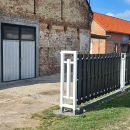 Nowoczesne ogrodzenie panelowe z czarnymi sztachetami i białymi słupkami na tle ceglanego budynku i budowy domu.