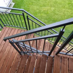 Zewnętrzne schody z drewnianymi stopniami i czarną metalową balustradą, widok z góry, z elementami ogrodu w tle.