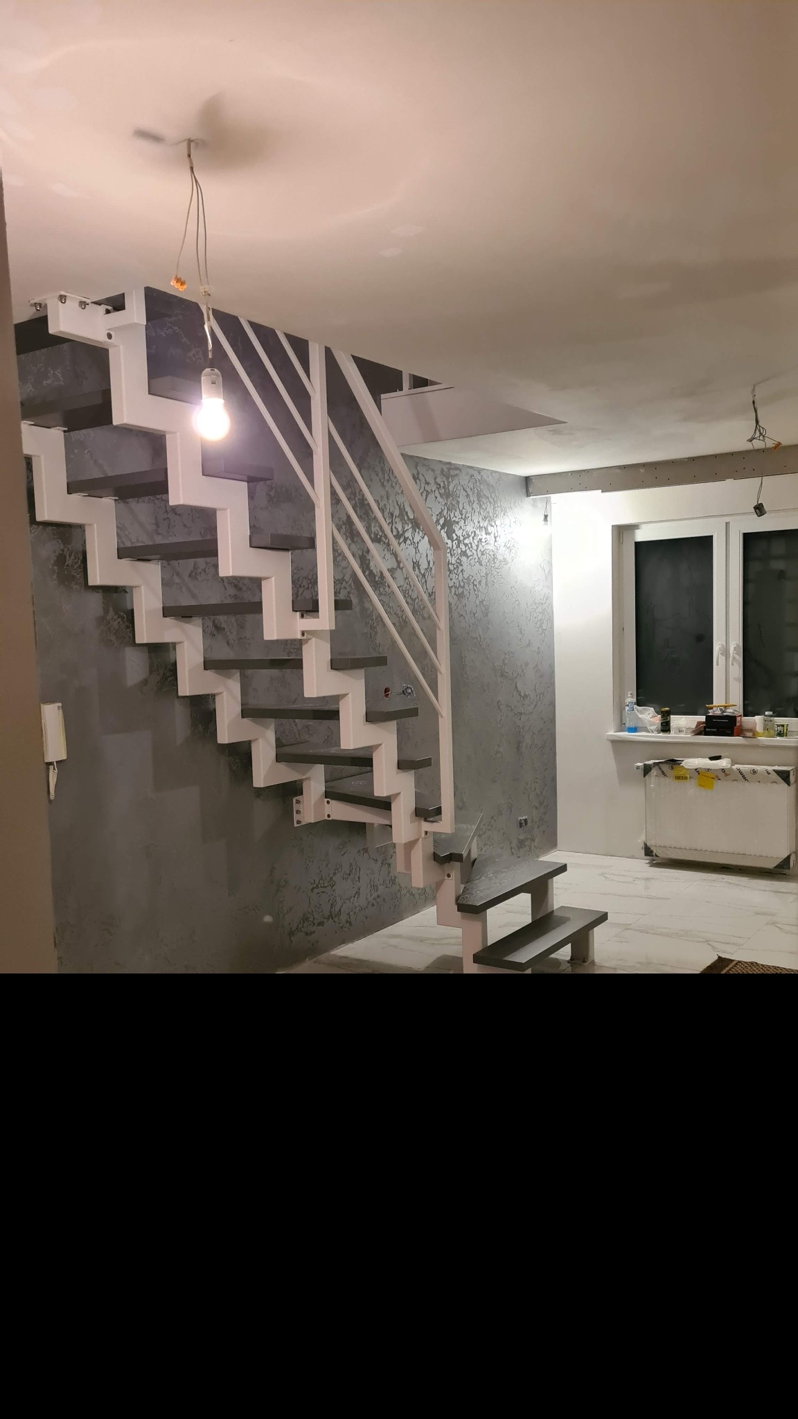 Nowoczesne schody metalowe z białą konstrukcją i szarymi stopniami w jasnym wnętrzu z ozdobną ścianą i marmurową podłogą, widoczne niedokończone prace wykończeniowe.