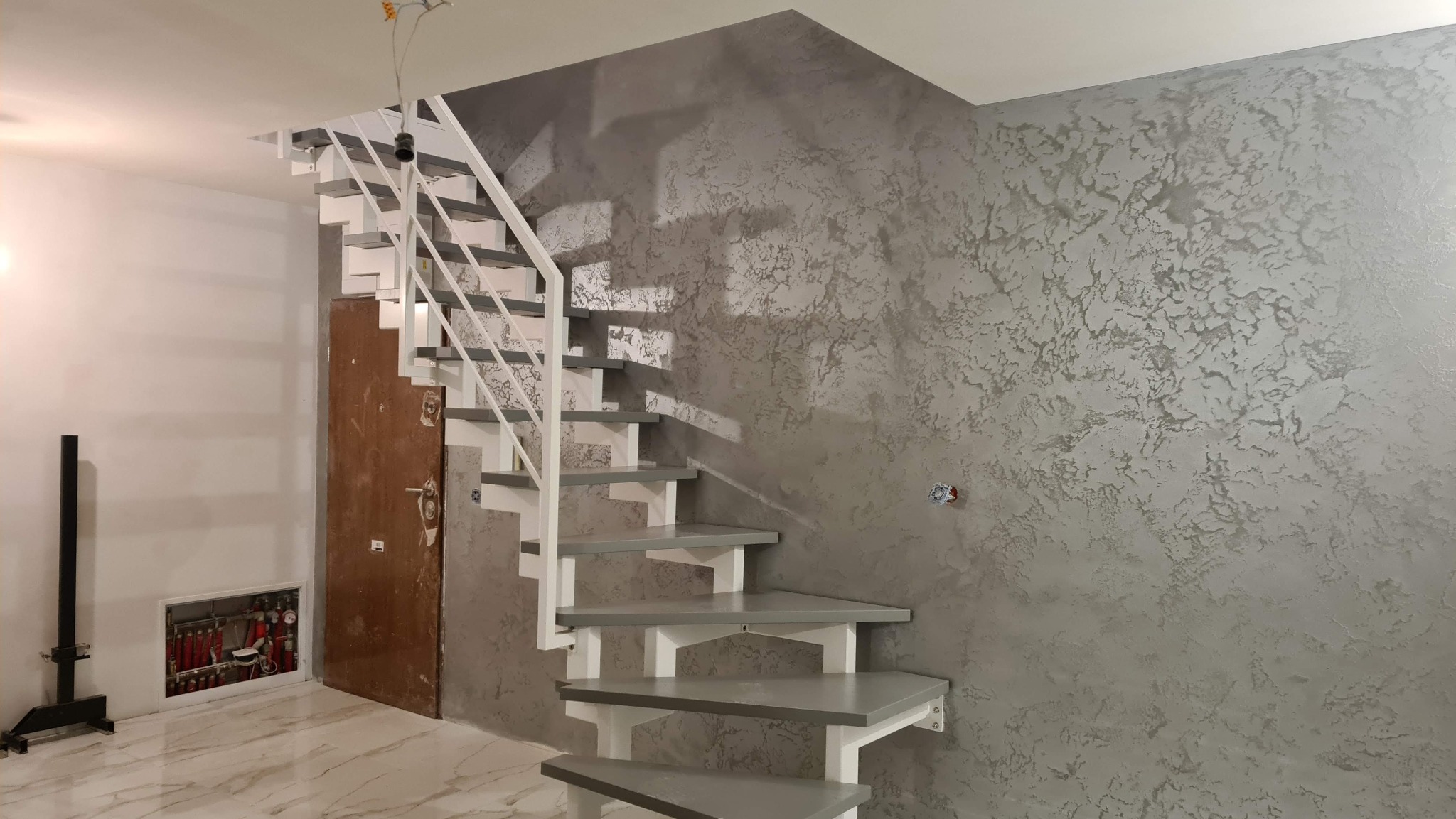Nowoczesne schody metalowe z białymi poręczami i szarymi stopniami, zamontowane w pomieszczeniu z dekoracyjną szarą ścianą i marmurową podłogą.