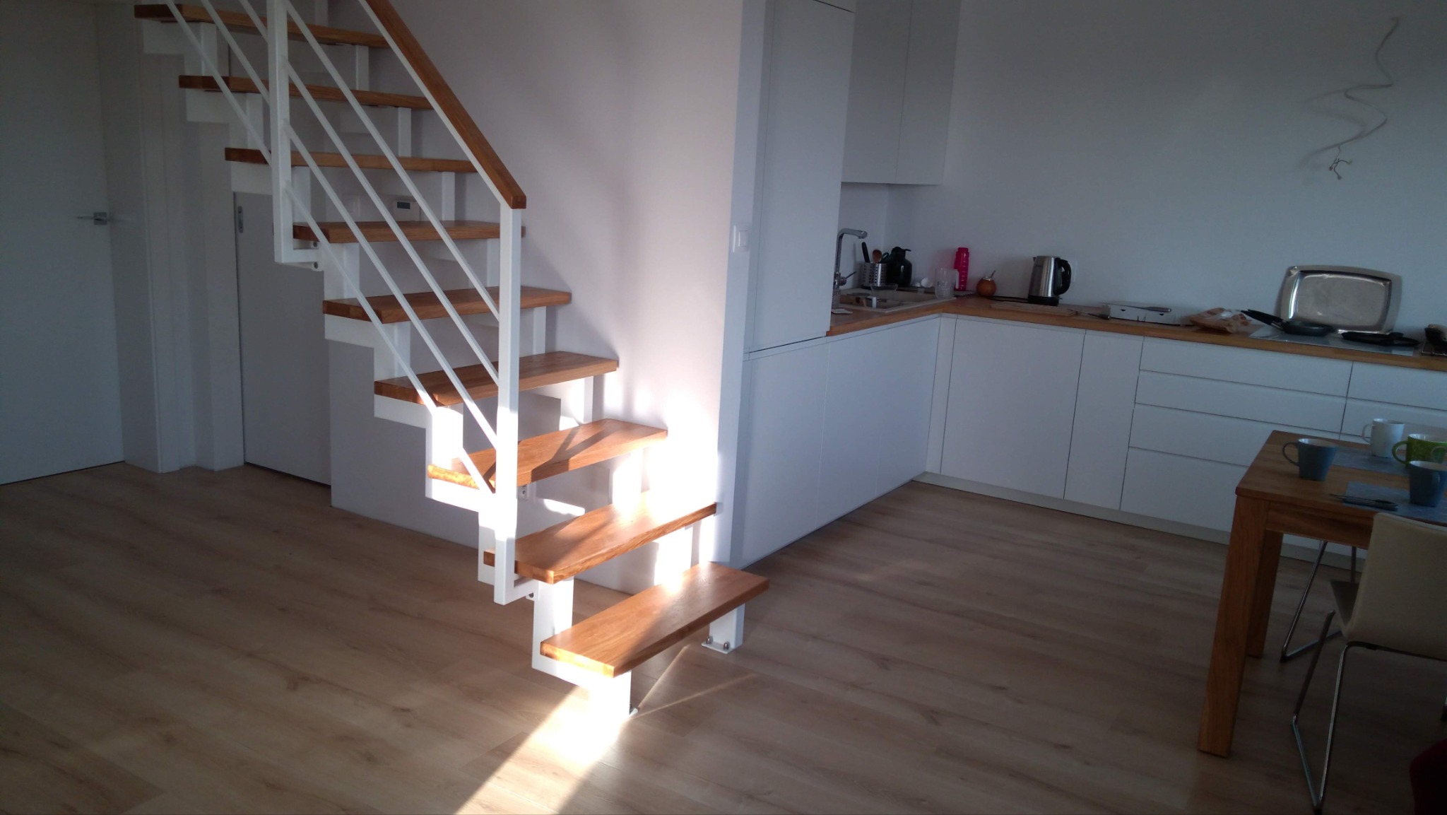 Nowoczesne schody wewnętrzne z drewnianymi stopniami i białą metalową konstrukcją w jasnym wnętrzu z widoczną kuchnią.