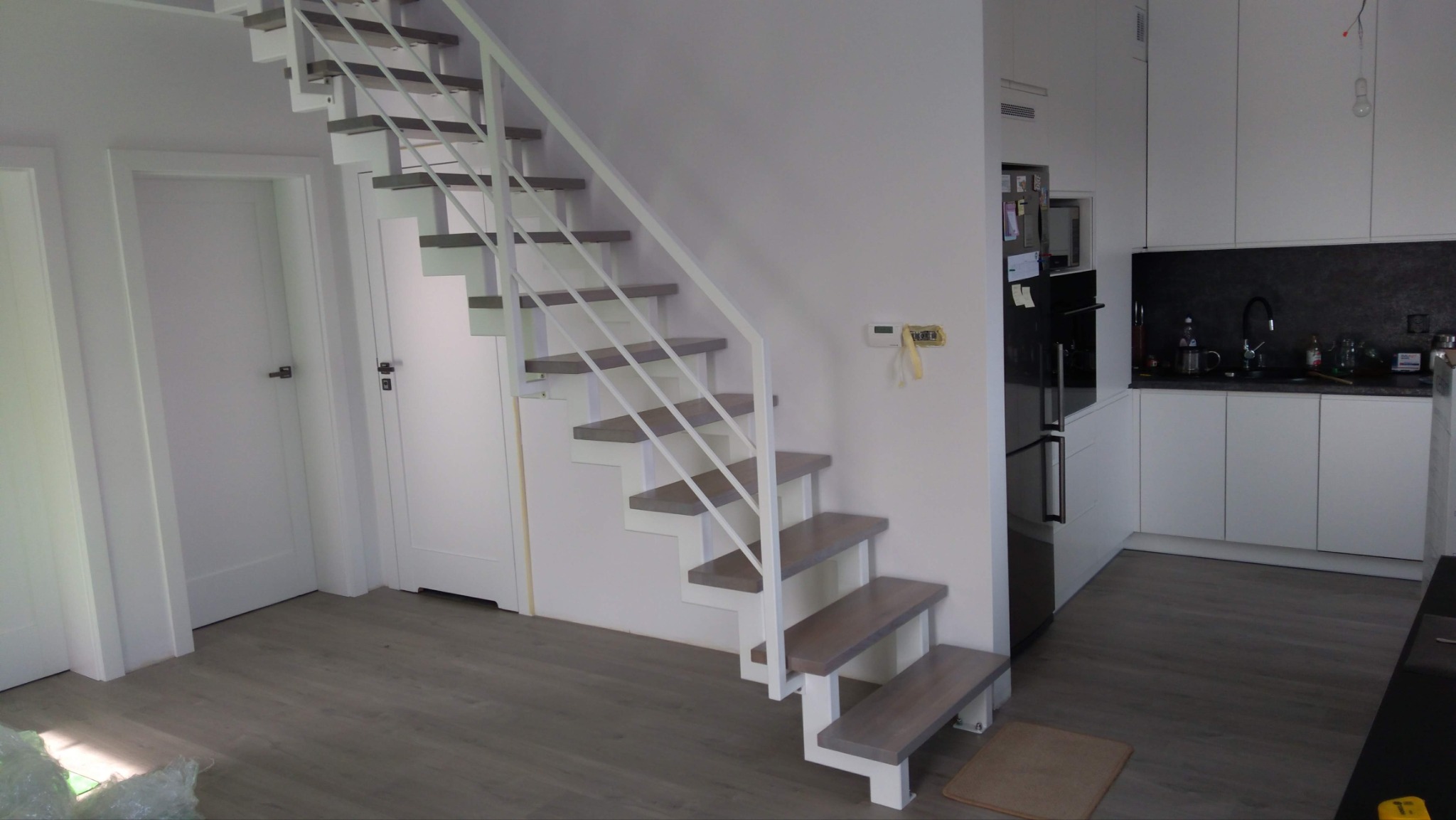 Metalowe schody z białymi, minimalistycznymi balustradami w domu w Skrzypniku, widoczne z dołu, prowadzące na wyższą kondygnację, z jasnym wnętrzem i fragmentem białej kuchni w tle.