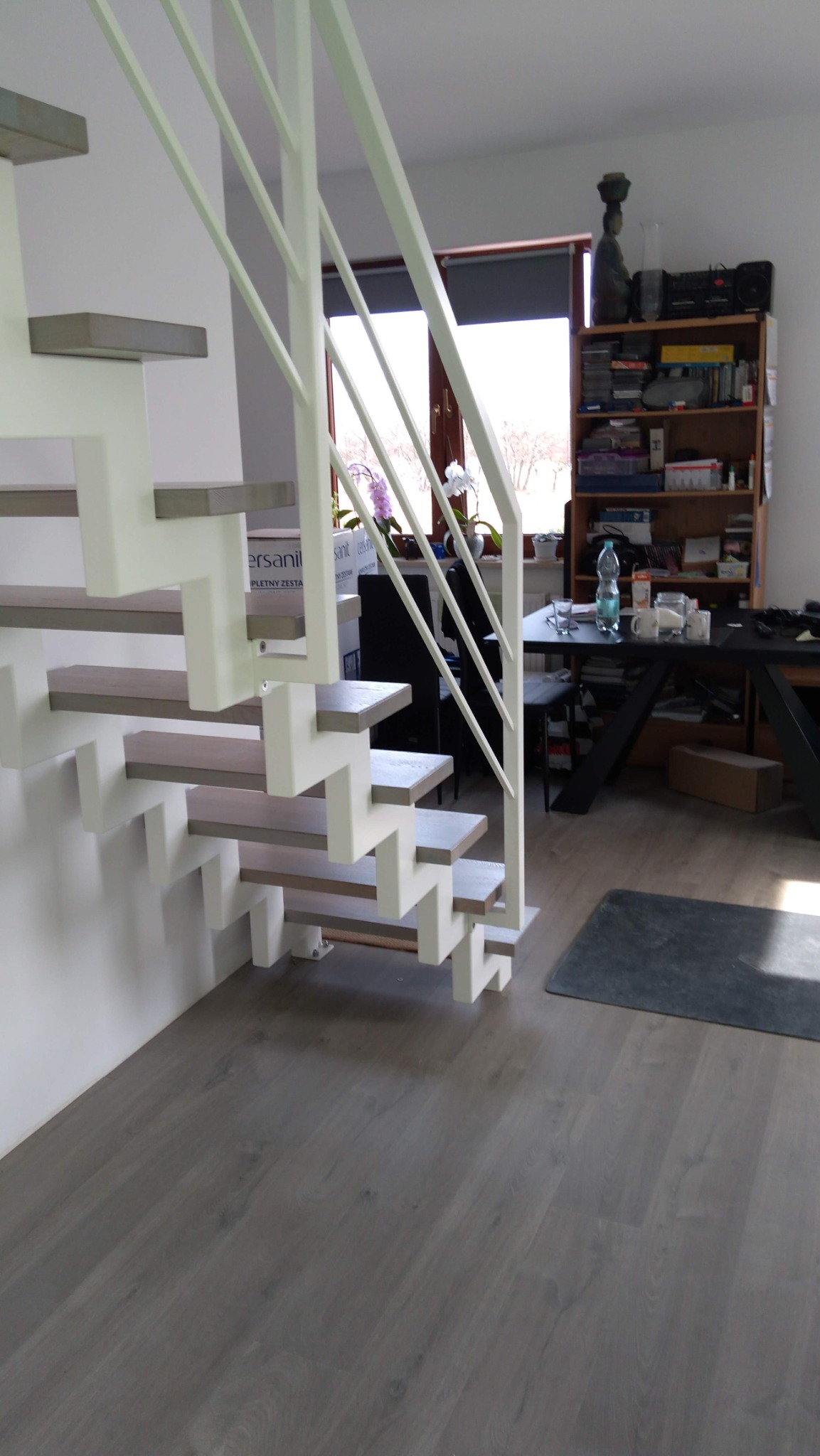 Nowoczesne schody z białymi wspornikami i szarymi stopniami, zabezpieczone białą, minimalistyczną balustradą, widoczne z dołu, w tle okno i regał z książkami.