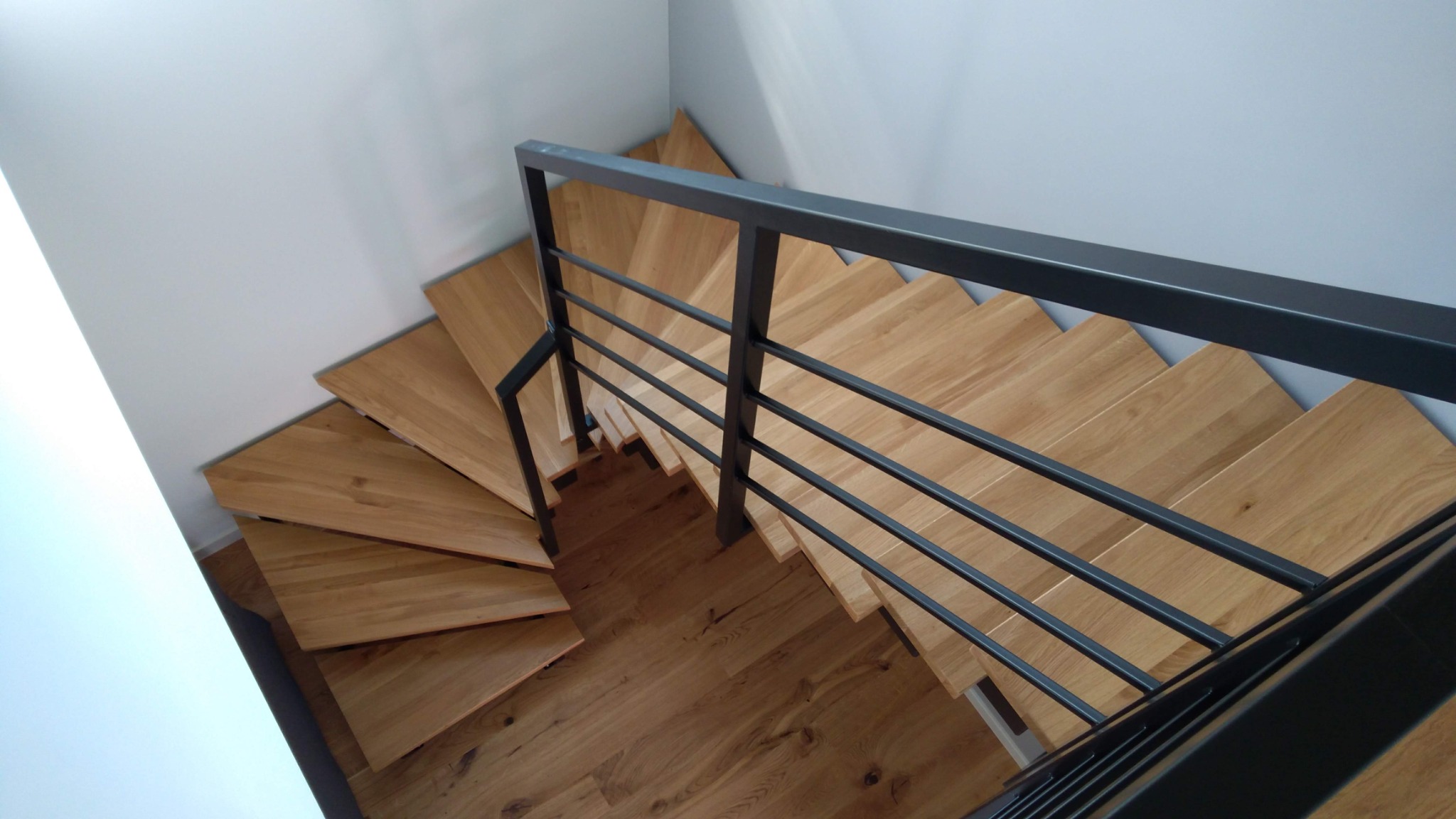 Kątowe ujęcie drewnianych schodów z czarną, metalową balustradą, widoczne z góry, z jasnymi ścianami w tle.