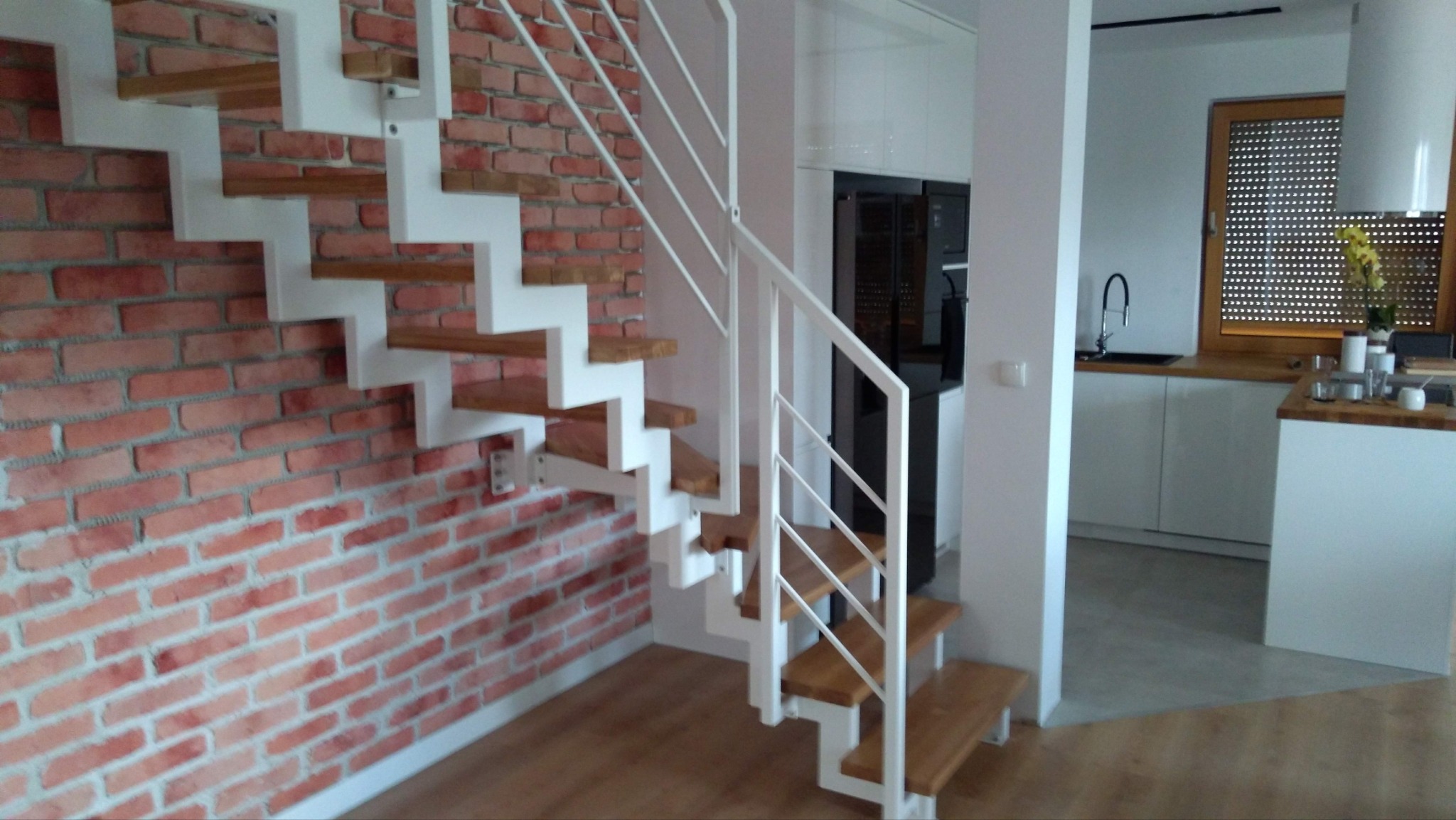 Nowoczesne schody z drewnianymi stopniami i białą metalową konstrukcją, zamontowane przy ścianie z czerwonej cegły, w tle widoczna kuchnia z białymi szafkami.