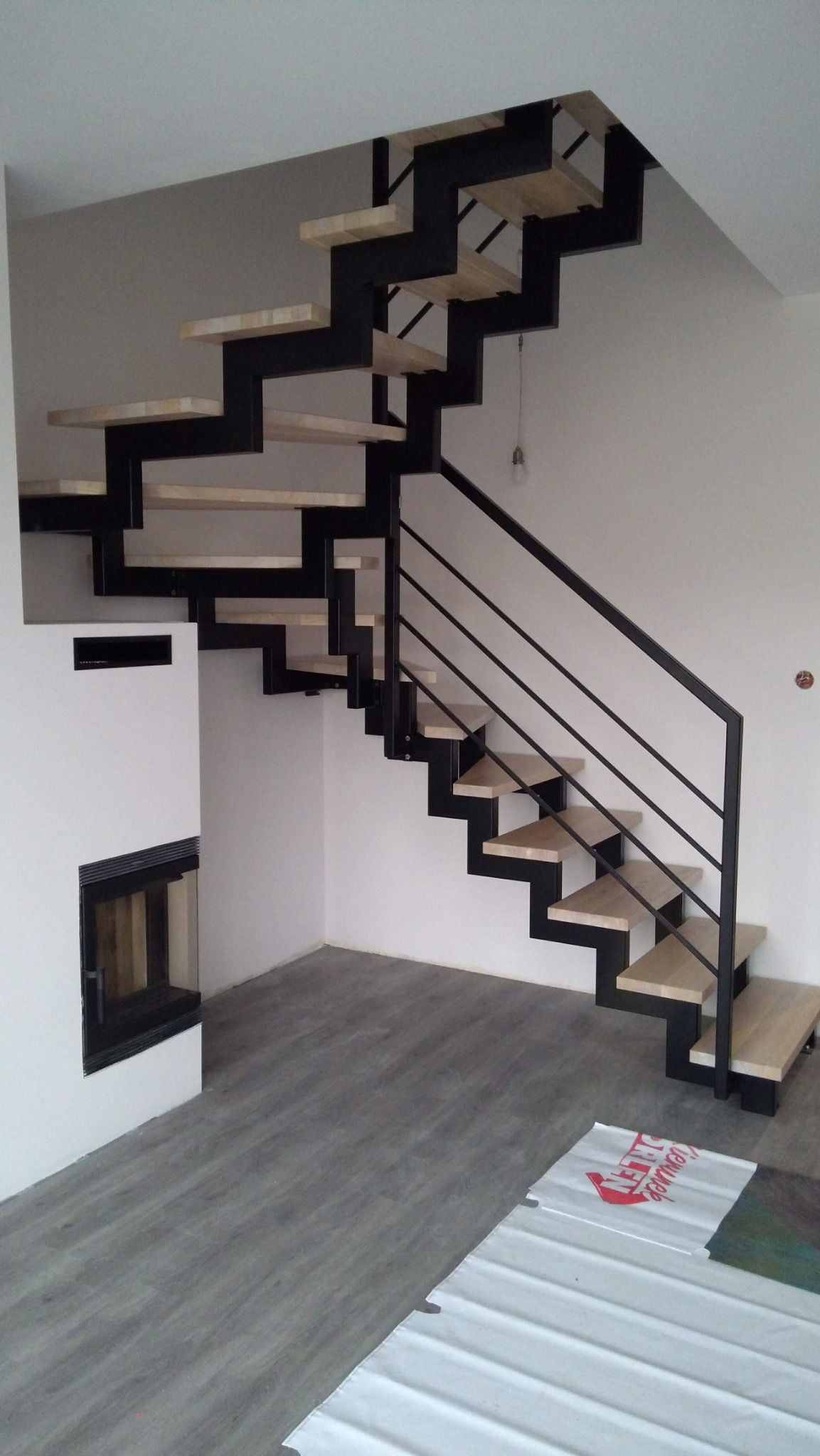Nowoczesne schody z czarną metalową konstrukcją i jasnymi drewnianymi stopniami, widoczne w jasnym wnętrzu z kominkiem i szarymi panelami podłogowymi, częściowo zakryte folią ochronną.