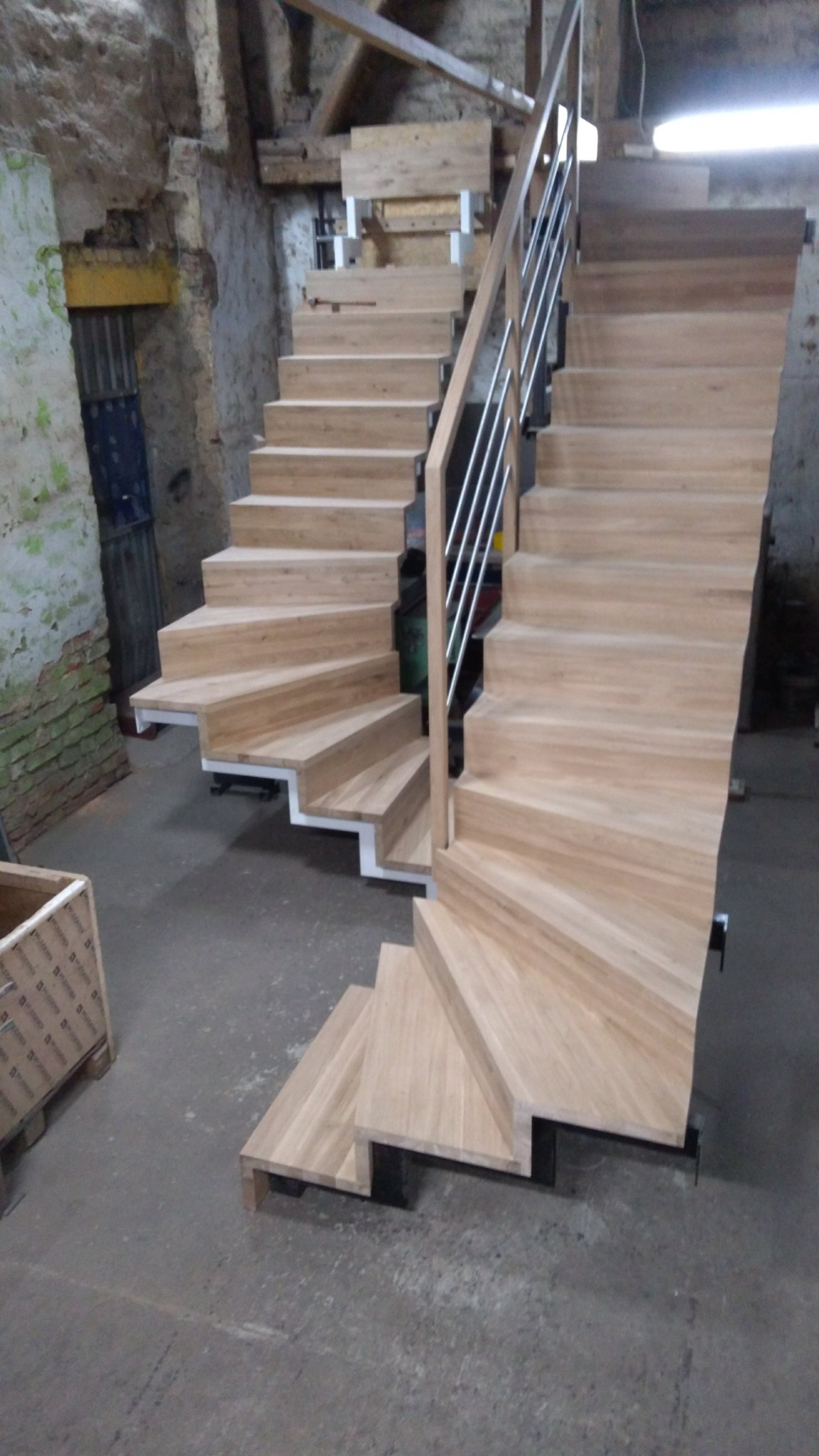 Drewniane schody dwubiegowe z metalową balustradą w surowym wnętrzu, widoczne detale konstrukcyjne i wykończenie drewna.