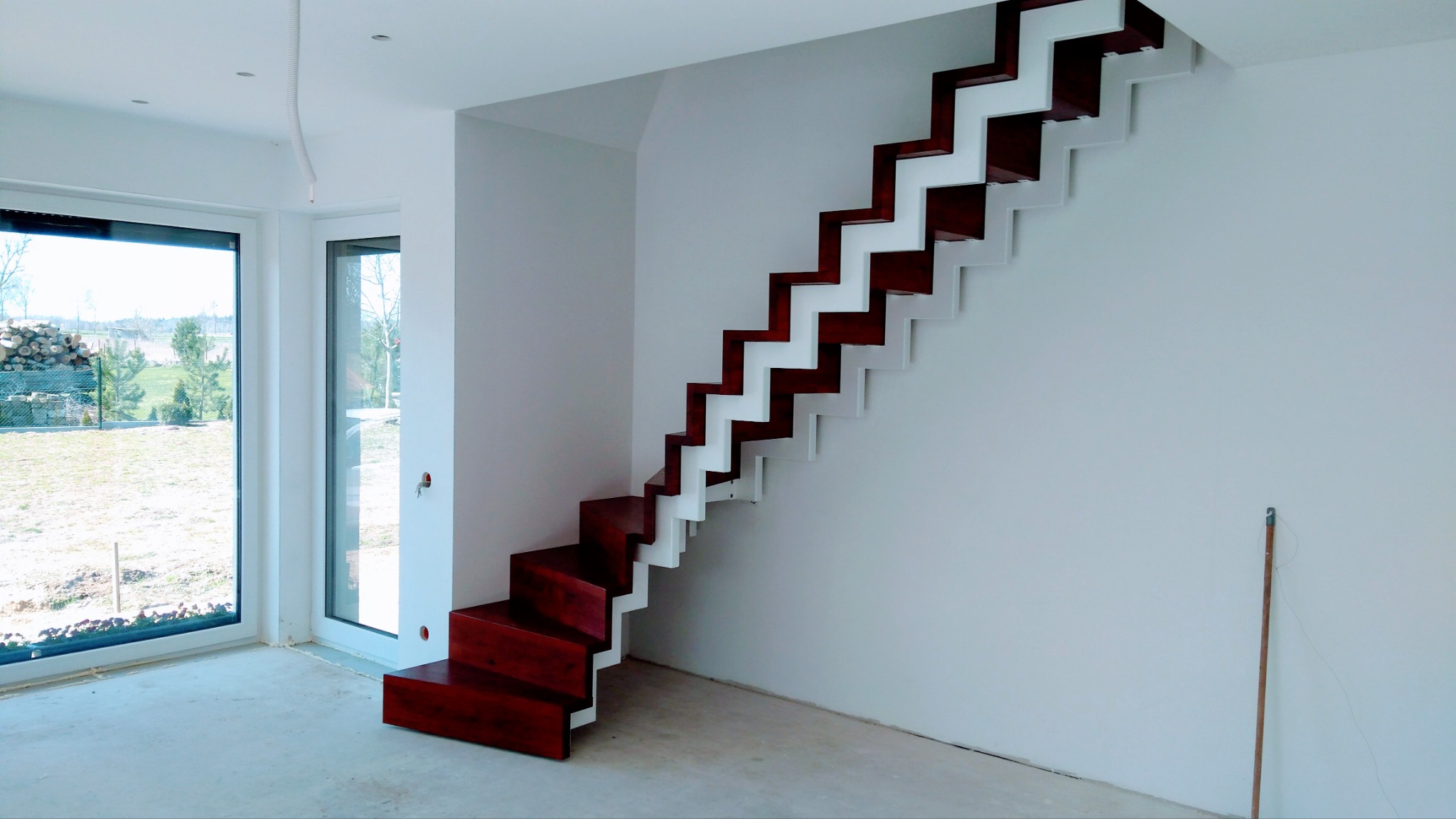 Nowoczesne, samonośne schody z ciemnego drewna i białymi wspornikami, widoczne na tle białych ścian i okna z widokiem na ogród w surowym stanie.