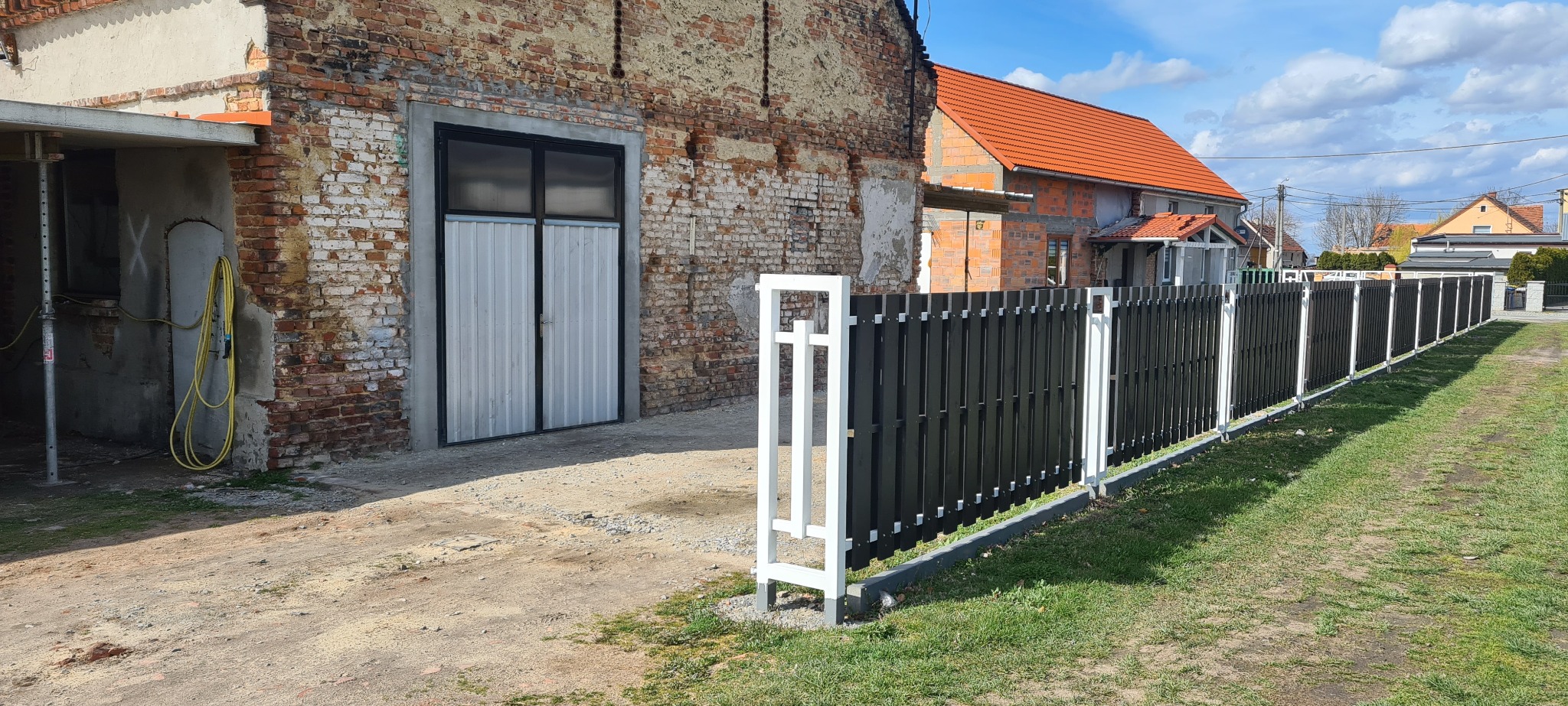 Nowoczesne ogrodzenie panelowe z czarnymi sztachetami i białymi słupkami na tle ceglanego budynku i budowy domu.