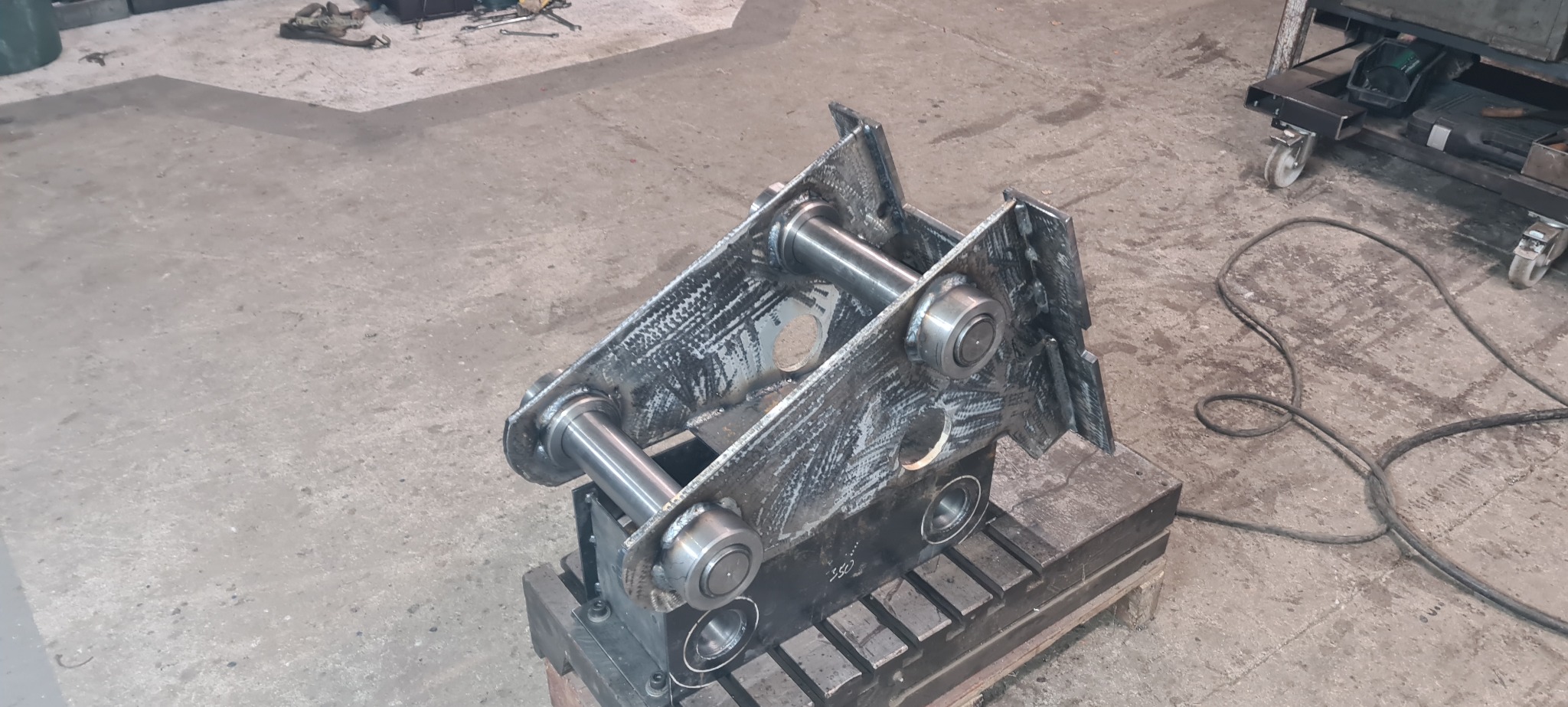 Spawana konstrukcja metalowa z otworami i cylindrycznymi elementami, umieszczona na metalowym stole warsztatowym, widoczne ślady po spawaniu.