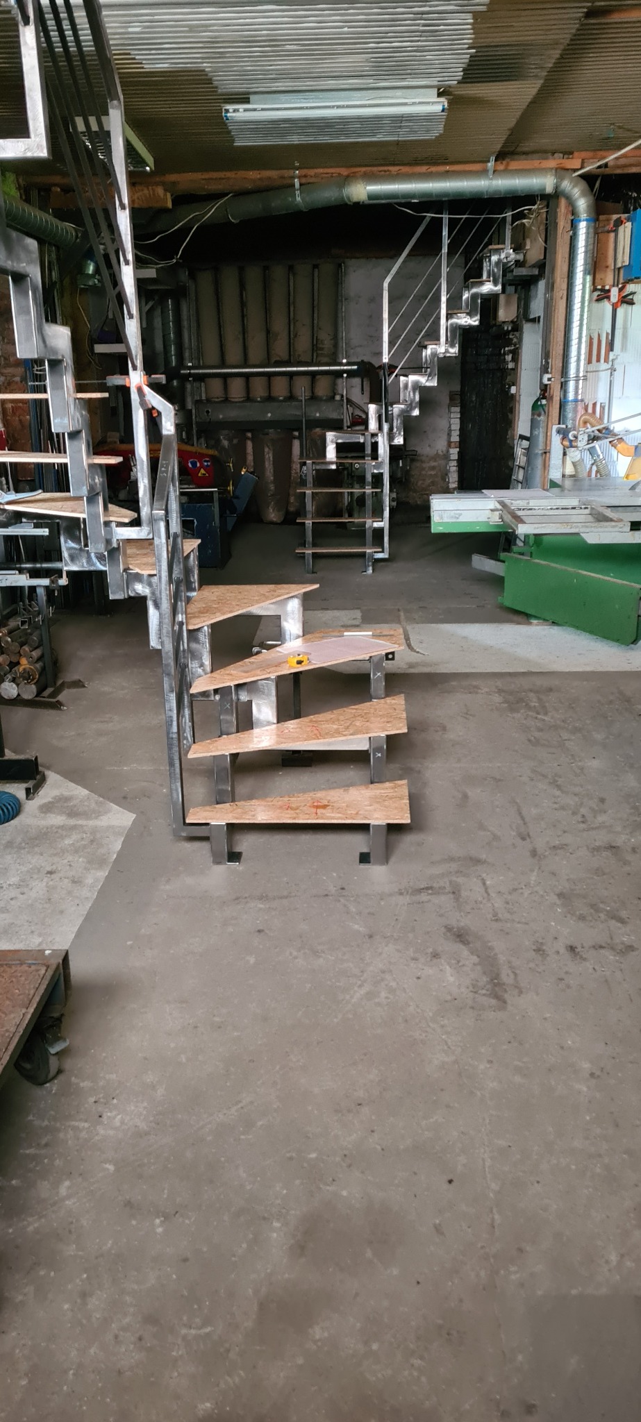 Metalowa konstrukcja schodów z drewnianymi stopniami w trakcie montażu w warsztacie, widoczne narzędzia pomiarowe na jednym ze stopni.