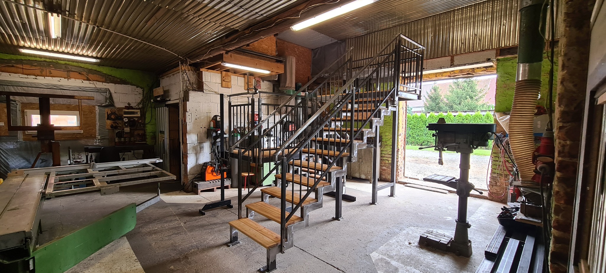 Metalowe schody z drewnianymi stopniami w warsztacie ślusarskim, widoczna spawarka, wiertarka stołowa i narzędzia.