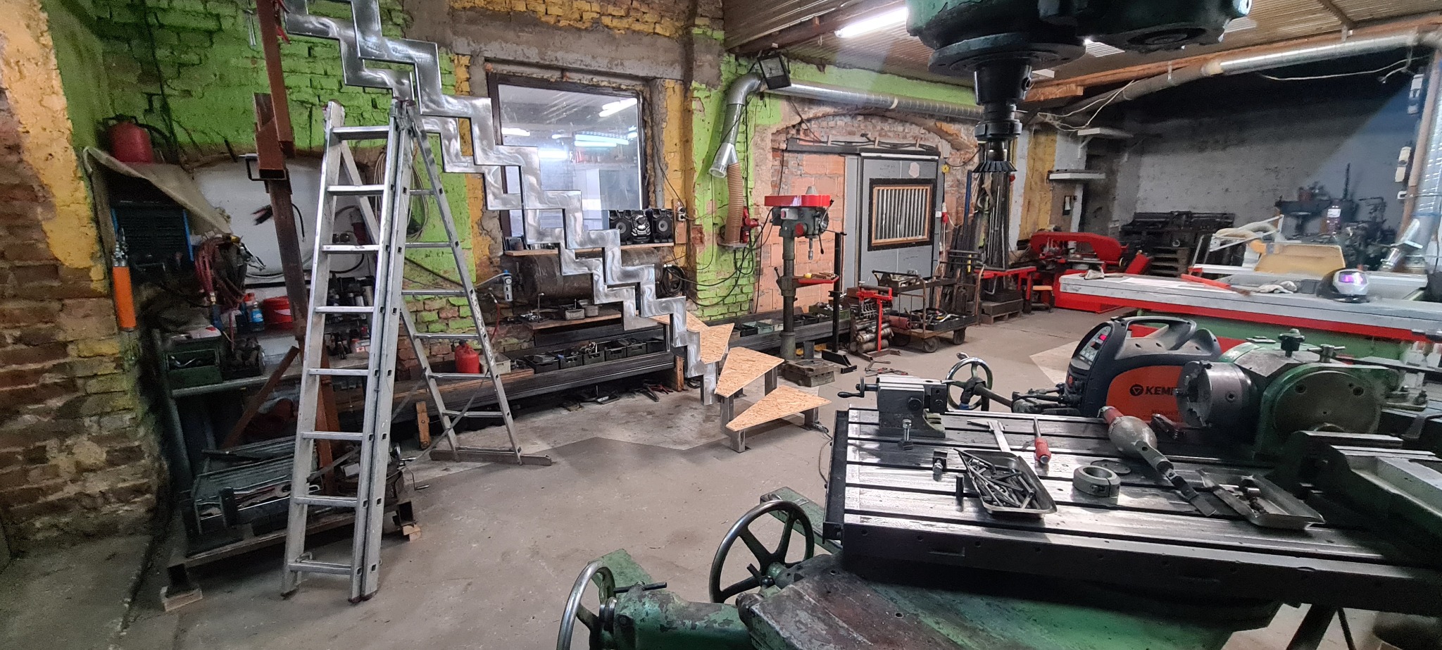 Warsztat z elementami metalowych schodów, drabina oparta o konstrukcję schodową, maszyny do obróbki metalu w tle ceglane ściany.