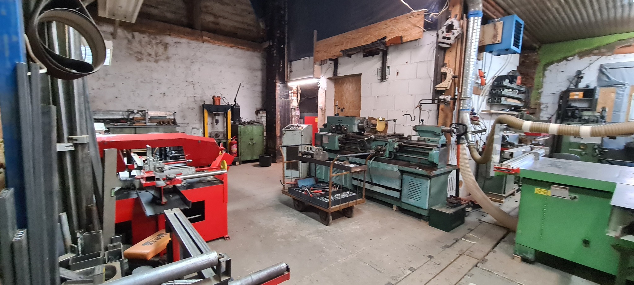 Warsztat ślusarski z maszynami do obróbki metalu, w tle widoczna piła taśmowa do metalu w kolorze czerwonym, tokarka, frezarka i inne narzędzia.