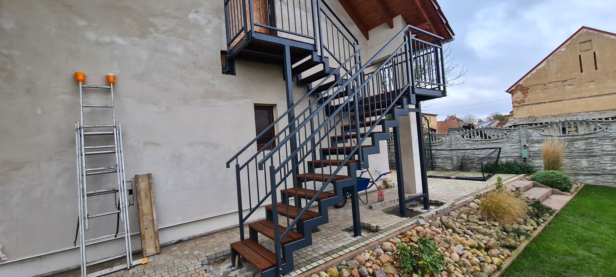 Zewnętrzne schody metalowe z drewnianymi stopniami prowadzące na balkon, widoczne narzędzia i drabina na tle budynku z dekoracyjnym wykończeniem.