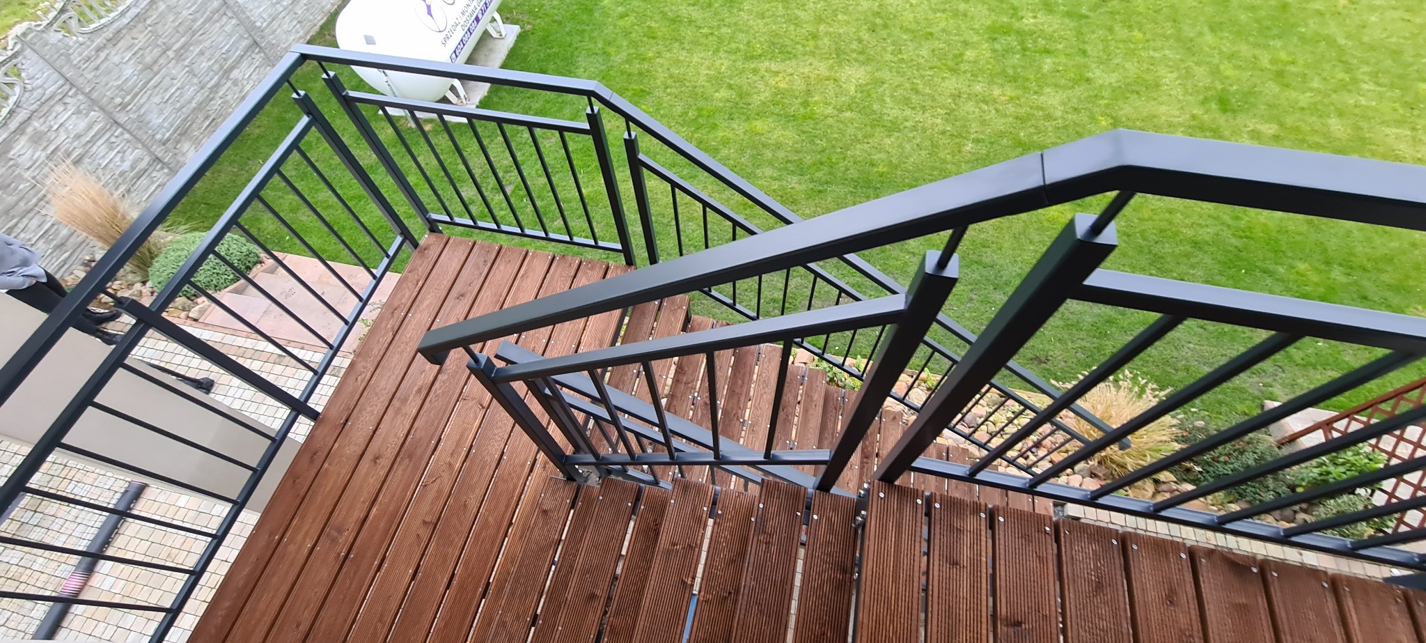 Zewnętrzne schody z drewnianymi stopniami i czarną metalową balustradą, widok z góry, z elementami ogrodu w tle.