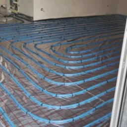 Niebieskie rury ogrzewania podłogowego ułożone spiralnie na folii z czerwoną kratką, widok z góry, przed wylewką, instalacja w surowym stanie.