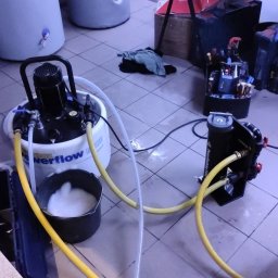 Usługi Remontowo - Budowlane Kamil Mościcki - Czyszczenie instalacji grzewczej za pomocą specjalistycznego urządzenia, widoczne węże, pompa i wiadro z pianą, obok skrzynka z narzędziami i zbiorniki.