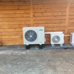 Trzy zewnętrzne jednostki klimatyzacyjne, w tym jedna marki York, ustawione na betonowej kostce brukowej przed drewnianą ścianą, z widocznym stosem drewna po lewej stronie.