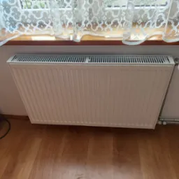 Biały grzejnik panelowy zamontowany pod oknem z koronkową firanką, widoczne rury doprowadzające i termostat.