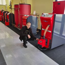 Usługi Remontowo - Budowlane Kamil Mościcki - Mężczyzna kuca obok kotła na paliwo stałe Atmos D 25 PX w showroomie, prezentując jego panel sterowania. W tle rzędy innych kotłów i zbiorników akumulacyjnych w kolorach czerwonym i szarym.