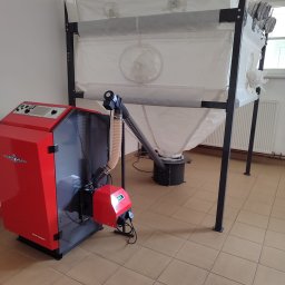 Usługi Remontowo - Budowlane Kamil Mościcki - Czerwono-szary piec na paliwo stałe z podajnikiem i zasobnikiem na pellet, widoczne elementy sterujące i podłączenie do podajnika, na brązowej podłodze z płytek.