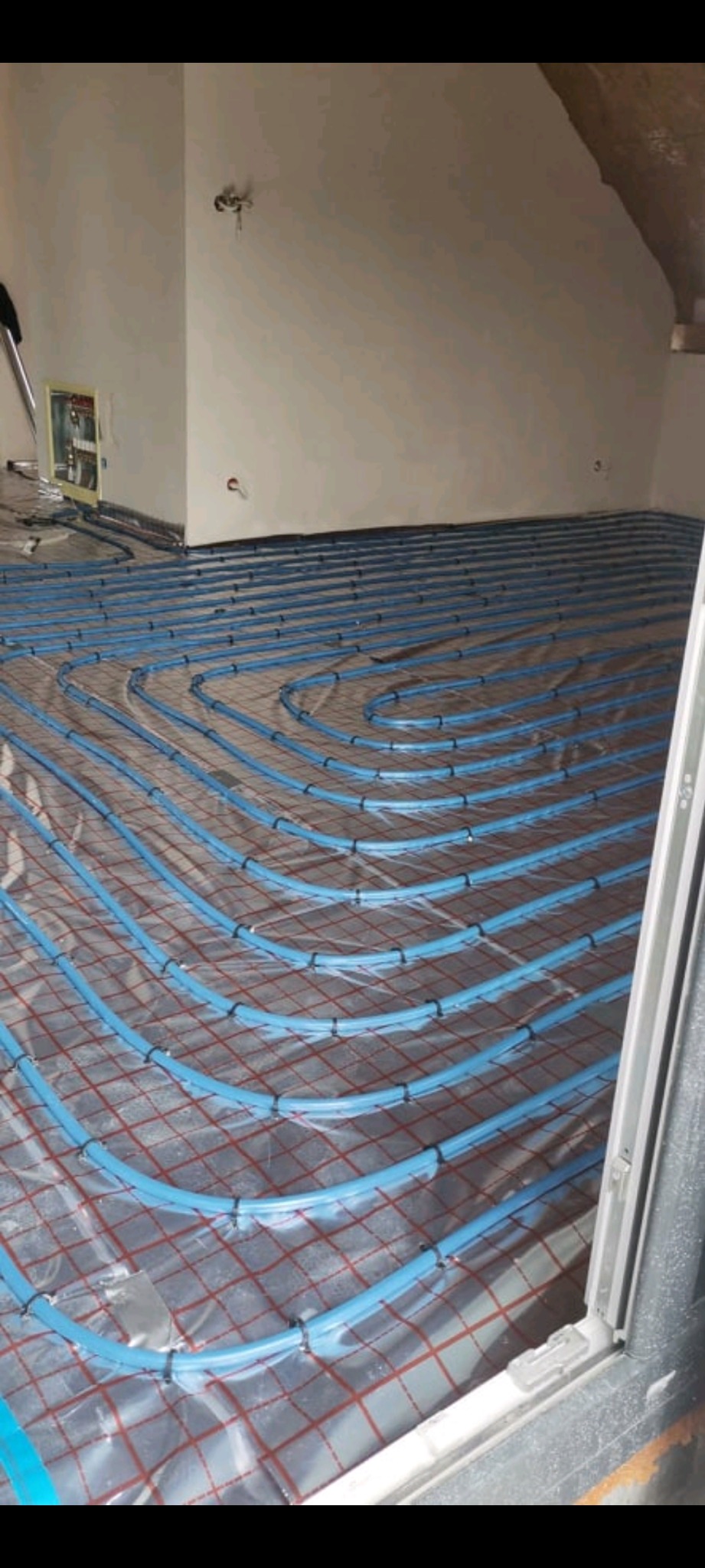 Niebieskie rury ogrzewania podłogowego ułożone spiralnie na folii z czerwoną kratką, widok z góry, przed wylewką, instalacja w surowym stanie.