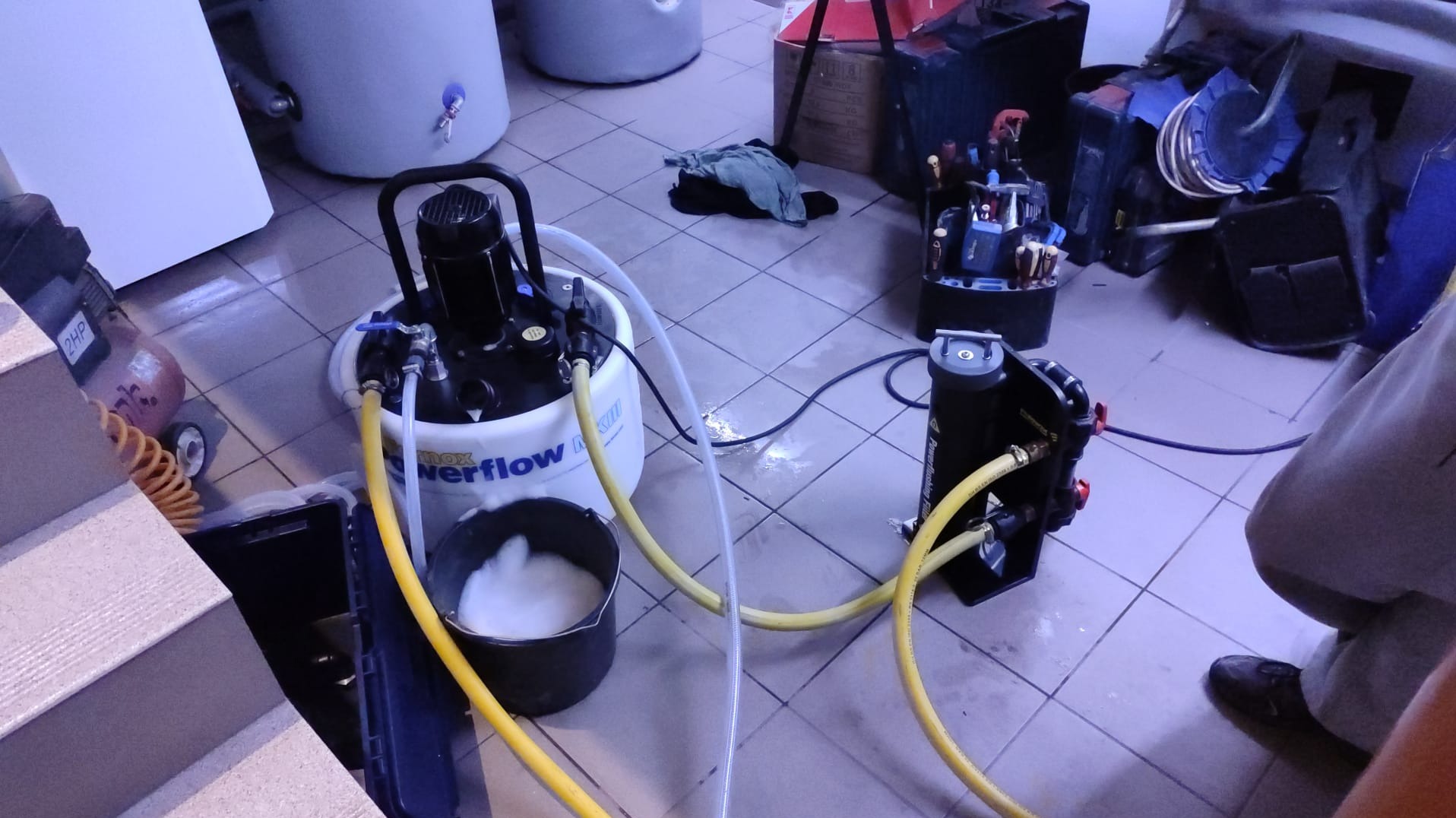 Czyszczenie instalacji grzewczej za pomocą specjalistycznego urządzenia, widoczne węże, pompa i wiadro z pianą, obok skrzynka z narzędziami i zbiorniki.