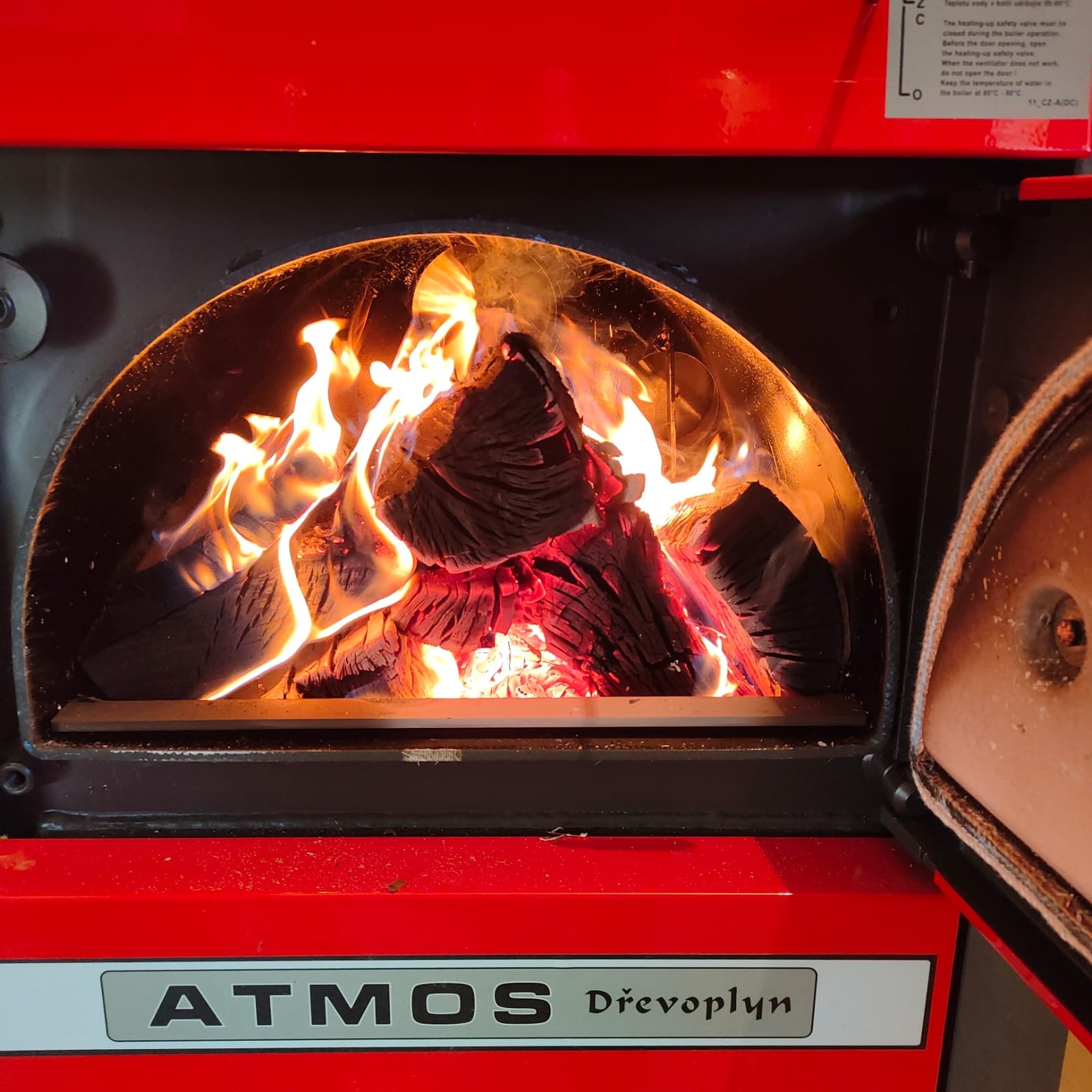 Czerwony piec na drewno marki Atmos Dřevoplyn z otwartymi drzwiczkami, wewnątrz płonące drewno i widoczne płomienie.