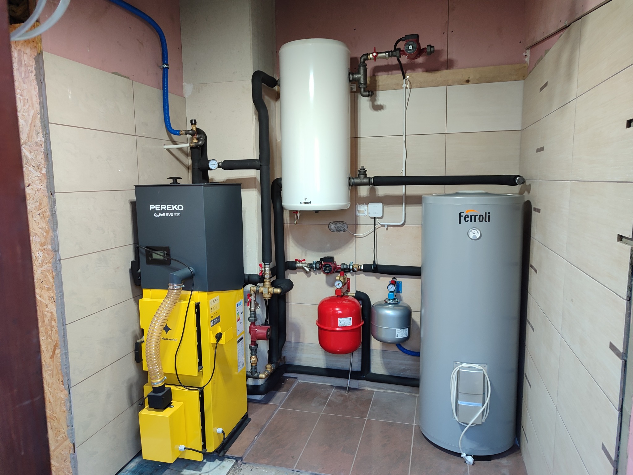 Kotłownia z piecem Pereko na pellet, zasobnikiem Galmet i zbiornikiem Ferroli, instalacja z rur w izolacji, płytki na ścianach i podłodze, widoczne elementy armatury i osprzętu.