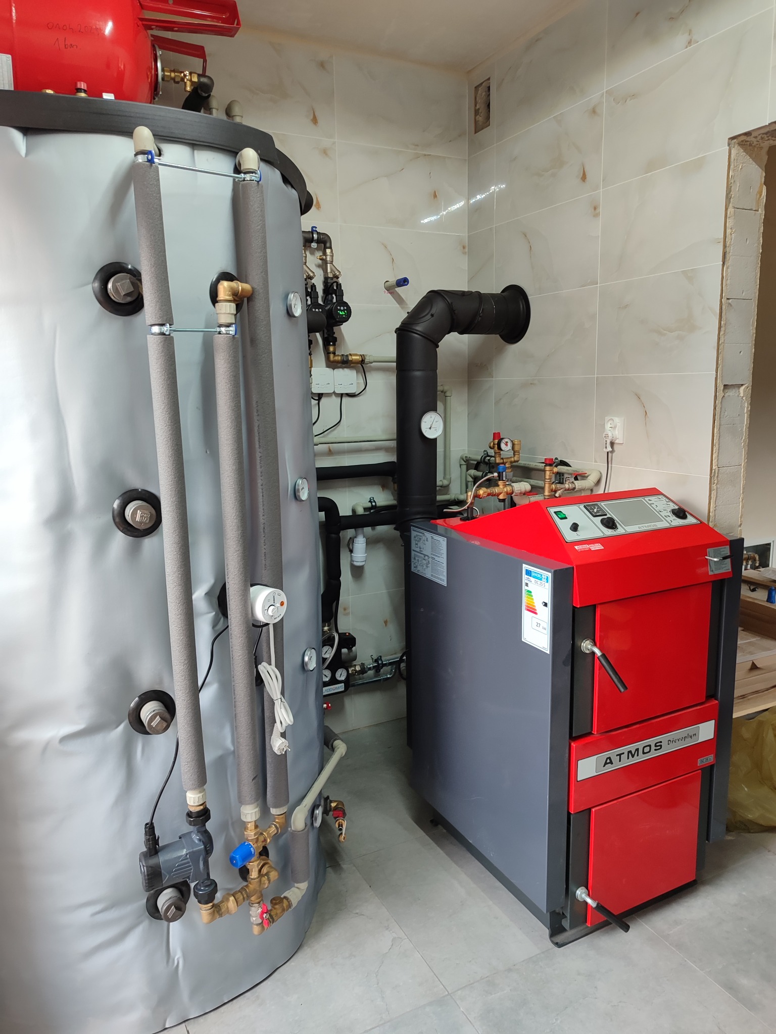 Nowoczesny piec na paliwo stałe Atmos DrevoPlyn w kotłowni z dużym, ocieplonym zasobnikiem akumulacyjnym i instalacją hydrauliczną z zaworami, manometrami i pompą obiegową, na tle płytek...