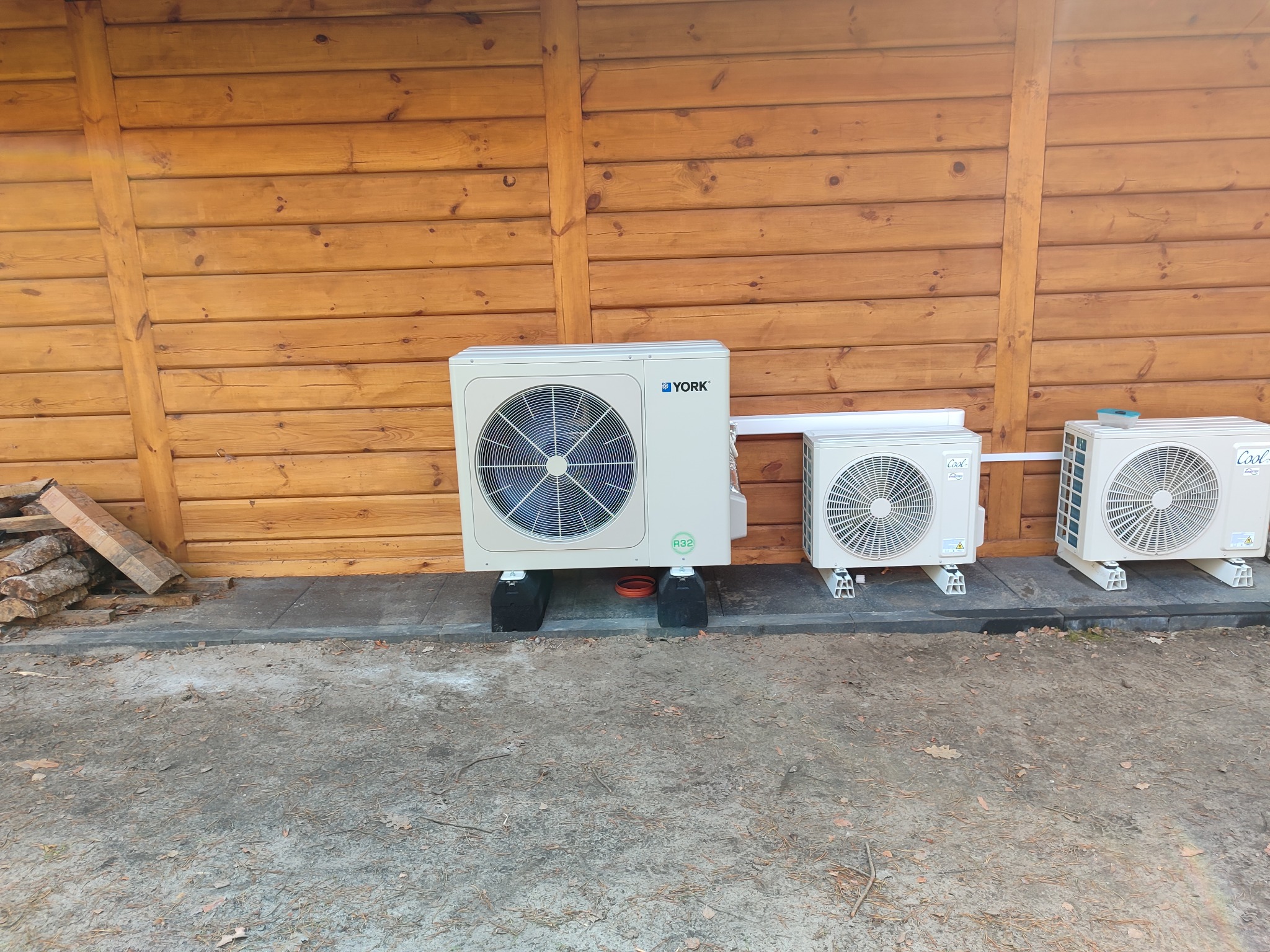 Trzy zewnętrzne jednostki klimatyzacyjne, w tym jedna marki York, ustawione na betonowej kostce brukowej przed drewnianą ścianą, z widocznym stosem drewna po lewej stronie.