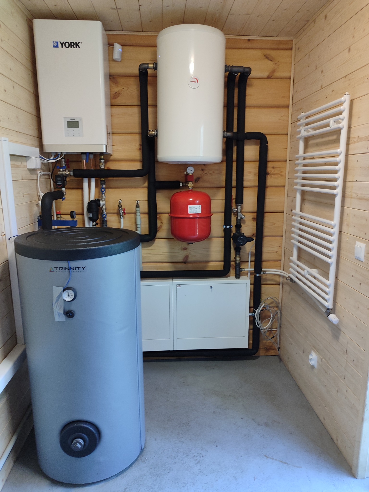 Kotłownia w drewnianym domku: piec gazowy York, zasobnik ciepłej wody, naczynie przeponowe w kolorze czerwonym oraz grzejnik drabinkowy.