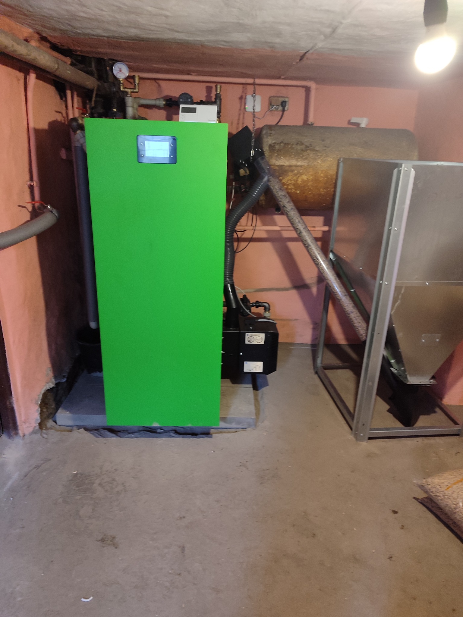 Zielony piec z podajnikiem na pellet w kotłowni, widoczne rury, zbiornik i worek z pelletem na podłodze.