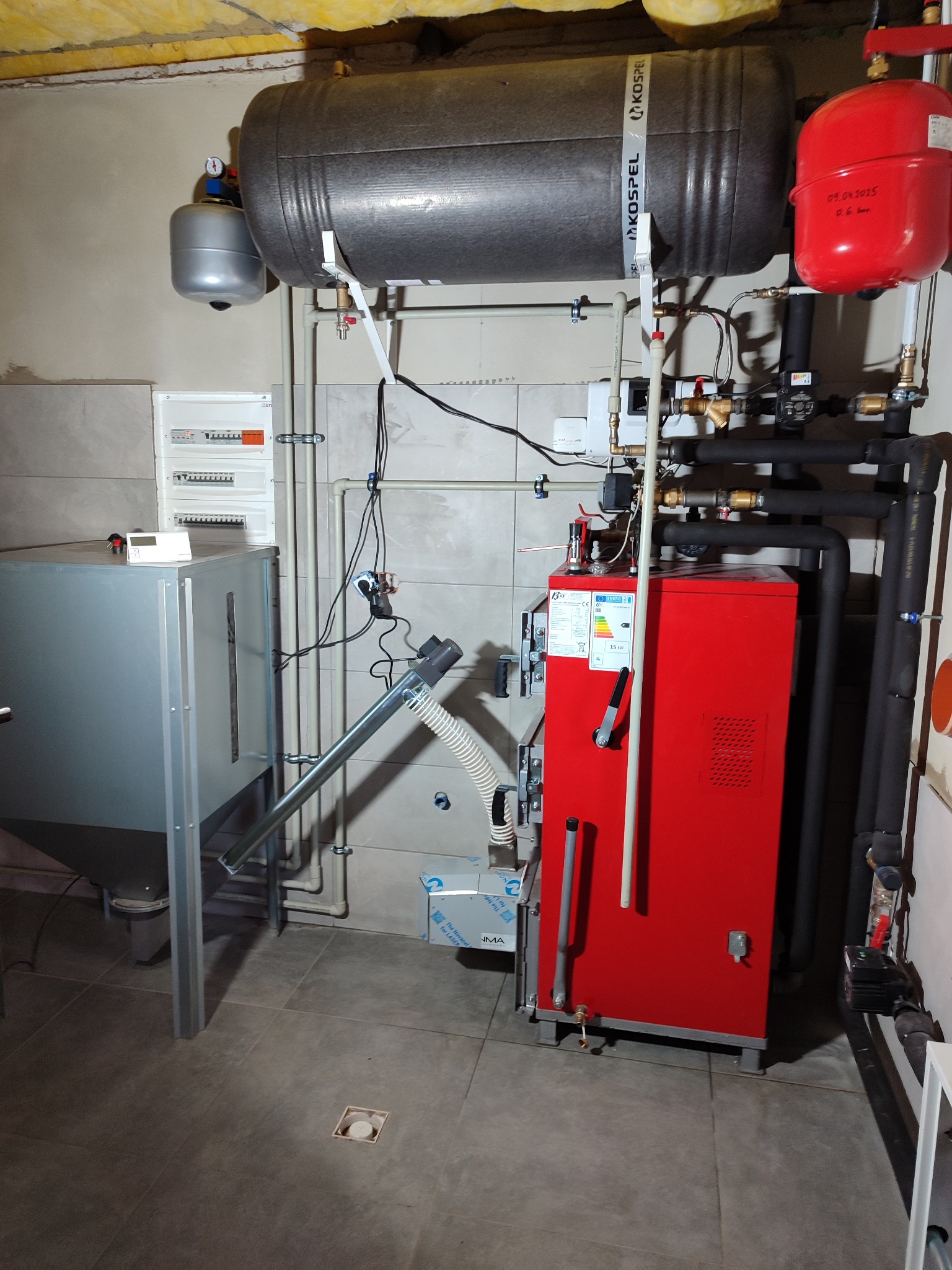 Czerwony piec centralnego ogrzewania z podajnikiem na ekogroszek i zbiornikiem buforowym, zainstalowany w kotłowni, widoczne rury, zawory, osprzęt i tablica elektryczna.