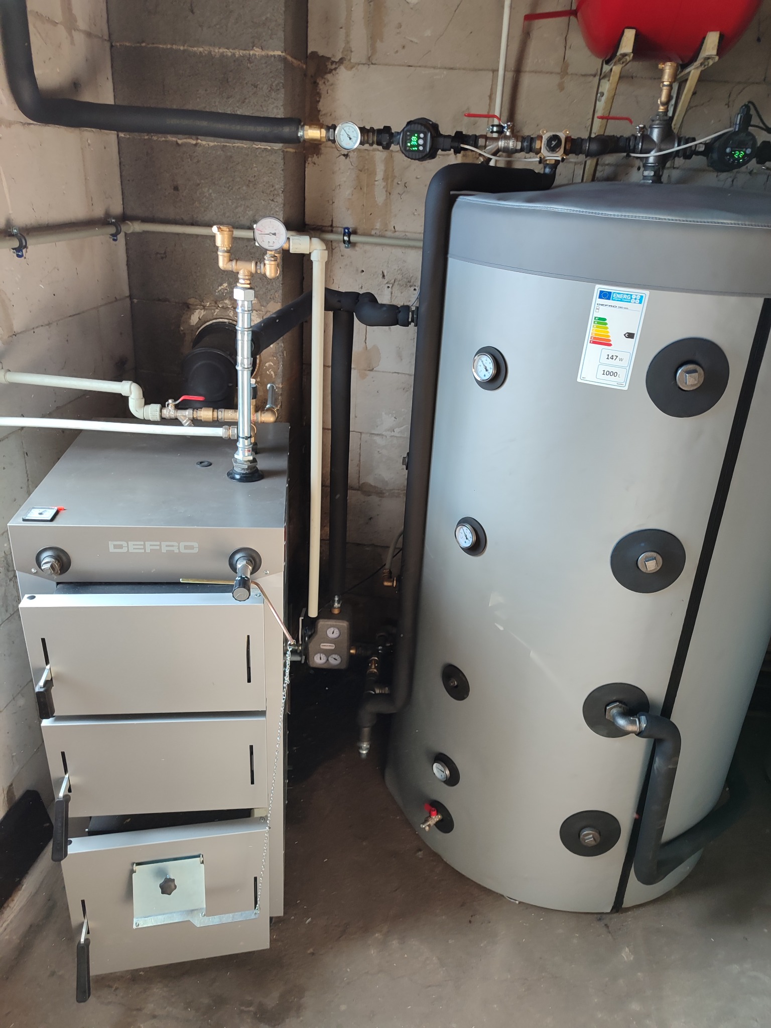 Szary piec Defro z otwartymi drzwiczkami obok szarego zbiornika akumulacyjnego z czarnymi zaślepkami, widoczne rury, manometry i instalacja w surowym pomieszczeniu.